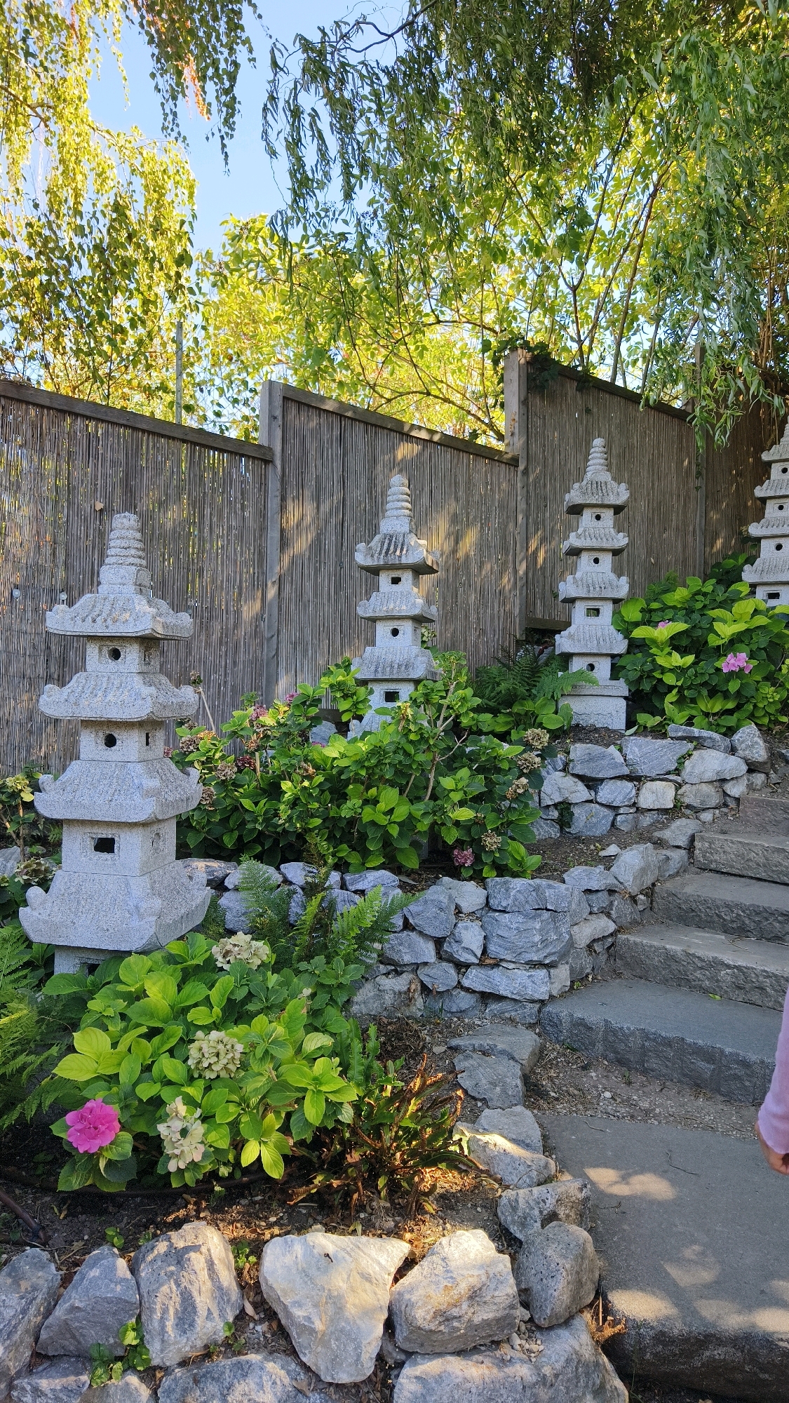 The image depicts a Japanese garden in Langenlois, Austria. In the foreground, a path made of large stone slabs leads upwards. To the left of the path, a series of tiered stone pagodas are arranged amongst lush green foliage and grey rocks. Several of the pagodas are visible, with the closest one being the largest. There are also some pink and white flowers interspersed with the plants. The background features a bamboo fence and trees with green leaves against a bright blue sky.