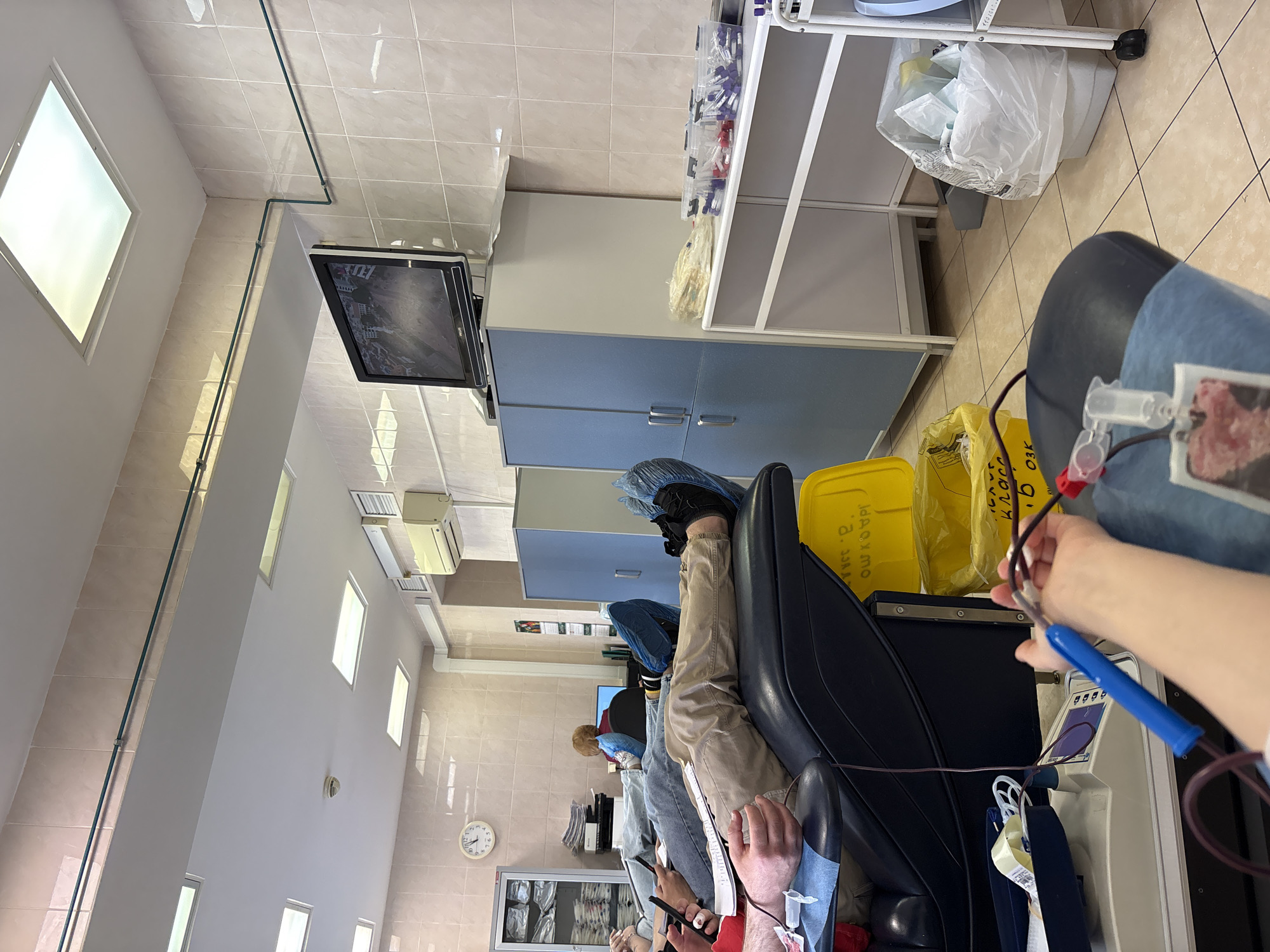 This image depicts an indoor scene within a blood donation or apheresis center in Saint Petersburg, Russia. The perspective is a low-angle shot, looking across a room equipped for medical procedures.
In the immediate foreground, a person is seated in a dark blue reclining medical chair, actively undergoing a blood donation or apheresis procedure. A needle is visible in their arm, connected by tubing to a collection system. Their hand holds a blue gripping device, commonly used by donors to facilitate blood flow. A white medical device with a digital display stands beside the chair.
Further back, at least two other individuals are visible, occupying similar reclining chairs, suggesting multiple donation stations. One person is notably wearing blue shoe covers, indicating a focus on maintaining a sterile or hygienic environment.
The room's interior features tiled floors and lower walls, transitioning to painted upper walls. Functional storage is provided by grey and blue cabinets along one wall, and a white metal shelving unit stocked with various medical supplies, including plastic vials. Several yellow medical waste bins are present, clearly labeled in Russian: "для опасных отходов" (for dangerous waste) and "Класс Б" (Class B), signifying hazardous medical waste.
High on a wall, a flat-screen television displays an unclear image, possibly a city scene. An air conditioning unit is mounted nearby. A round wall clock, showing the time around 11:25, is visible on a side wall. The setting is clean, orderly, and well-lit by rectangular ceiling lights, consistent with a clinical medical facility.