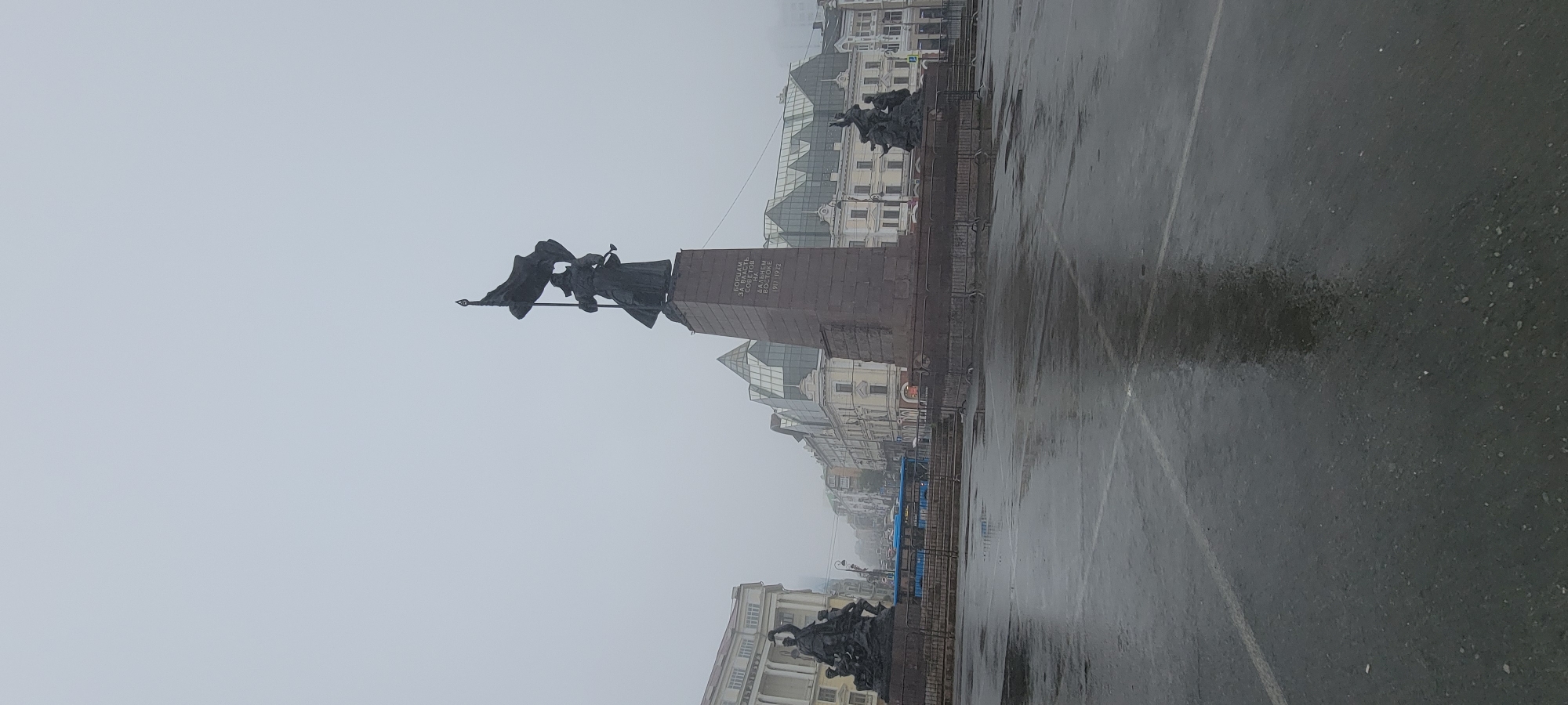The image depicts a monument in Vladivostok, Russia, on a foggy day. The monument, likely the "Borets za vlast' sovetov na Dal'nem Vostoke" (Fighters for Soviet Power in the Far East) monument, is centrally located and features a tall, tiered pedestal topped with a statue of a soldier holding a flag. Inscriptions are visible on the pedestal.
To the left of the monument, a group statue of figures is visible. To the right, another smaller statue group is present. The area around the monument appears to be an open plaza or square, with wet pavement reflecting the overcast sky, suggesting it has recently rained or is still raining. White painted lines are visible on the pavement, indicating parking or traffic lanes.
In the background, several buildings line the street, some appearing to be older architectural styles with ornate facades, while others have more modern glass structures. A blue bus is seen driving on the street behind the monument. The overall atmosphere is somber due to the fog and wet conditions.