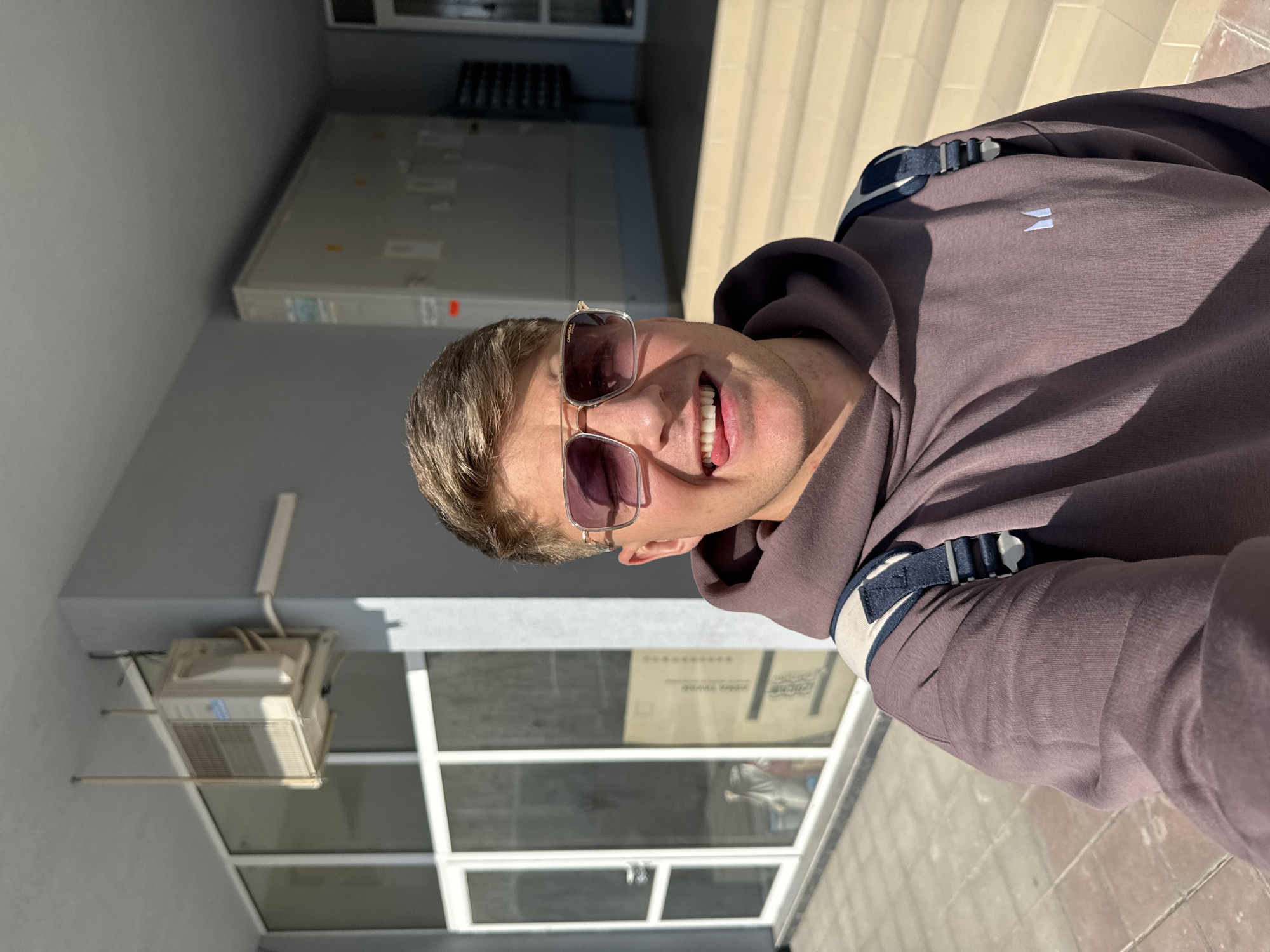 A medium shot depicts a fair-skinned male smiling at the camera. He wears sunglasses and a mauve hoodie with a small white logo. A backpack strap is visible over his shoulder. The background includes a building facade with a grey wall, white-framed windows, and an air conditioning unit. To the right are steps, suggesting an entrance. The location is outdoors, likely Plovdiv, Bulgaria, based on the image metadata.