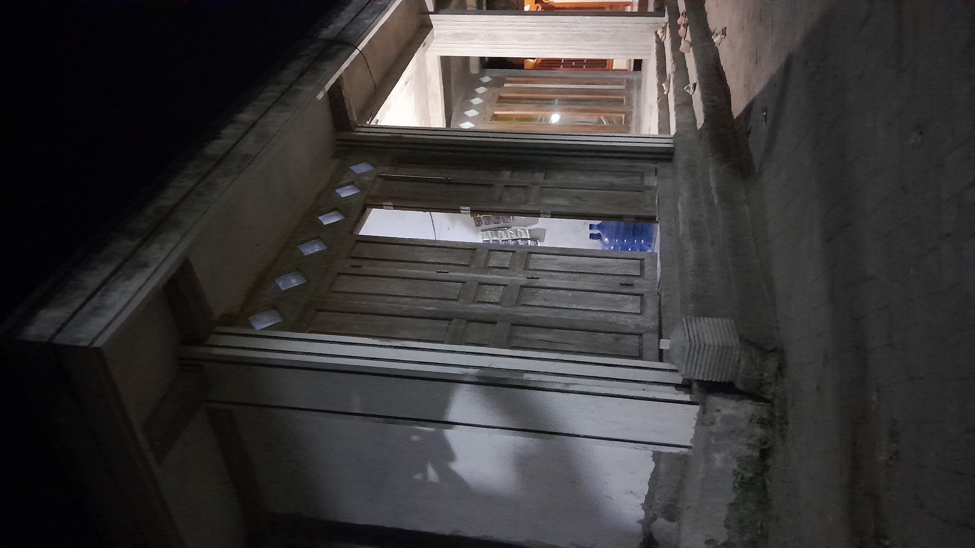 The image depicts an outdoor scene at night, likely in a commercial or residential area, with a focus on the entrance of a building. The building appears to be constructed of concrete and wood, with a large double wooden door that is slightly ajar, revealing a dimly lit interior. Two large blue water cooler bottles are visible just inside the doorway. Above the door, there are small, diamond-shaped windows that allow a faint light to escape. The building has a covered walkway or porch extending outwards, supported by concrete columns. In the foreground, a concrete pathway or sidewalk is visible, with some debris scattered on it. The ground is dark and appears to be paved. To the left of the entrance, a concrete wall with vertical lines is prominent, casting a shadow. To the right of the entrance, steps lead up to another part of the building, with several pairs of sandals or shoes placed on the steps. The overall lighting suggests it is nighttime, with artificial lights from within the building and possibly from streetlights or other sources. The location is identified as Sumberarum, Indonesia.