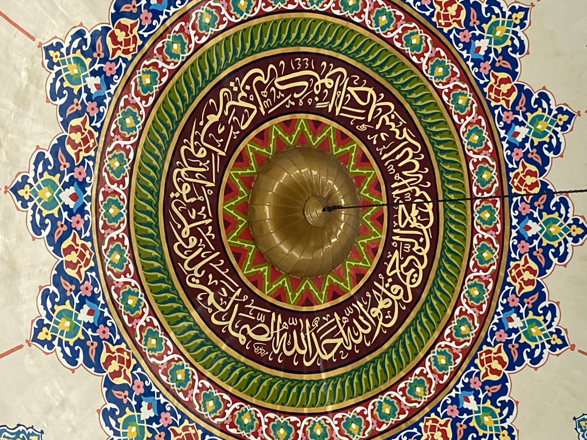 This is a close-up, overhead shot of a decorative ceiling in what appears to be a mosque. The central element is a golden, ribbed dome or finial, from which a dark cord hangs downwards. This central feature is surrounded by concentric circles of intricate geometric and calligraphic designs.
The innermost ring features a starburst pattern in red and green. Surrounding this is a wide band of dark red adorned with Arabic calligraphy in gold. Next, a thick green band with a wave-like pattern is visible, bordered by thinner gold rings. The outermost decorative layer is a richly colored border featuring blue, red, green, yellow, and pink floral and geometric motifs, set against a beige background.
The image does not contain any people, and the lighting suggests it was taken indoors, possibly with flash photography given the detail and color saturation. The style of decoration is characteristic of Islamic art, often found in mosques.
While the image itself doesn't provide specific location details, it is important to note that the city of Lhokseumawe, Indonesia, has several notable mosques, such as the Islamic Center Mosque of Lhokseumawe and the Baiturrahman Grand Mosque. These locations often feature similar decorative elements in their architecture, particularly on ceilings and domes, to enhance the spiritual and aesthetic ambiance for worshippers.