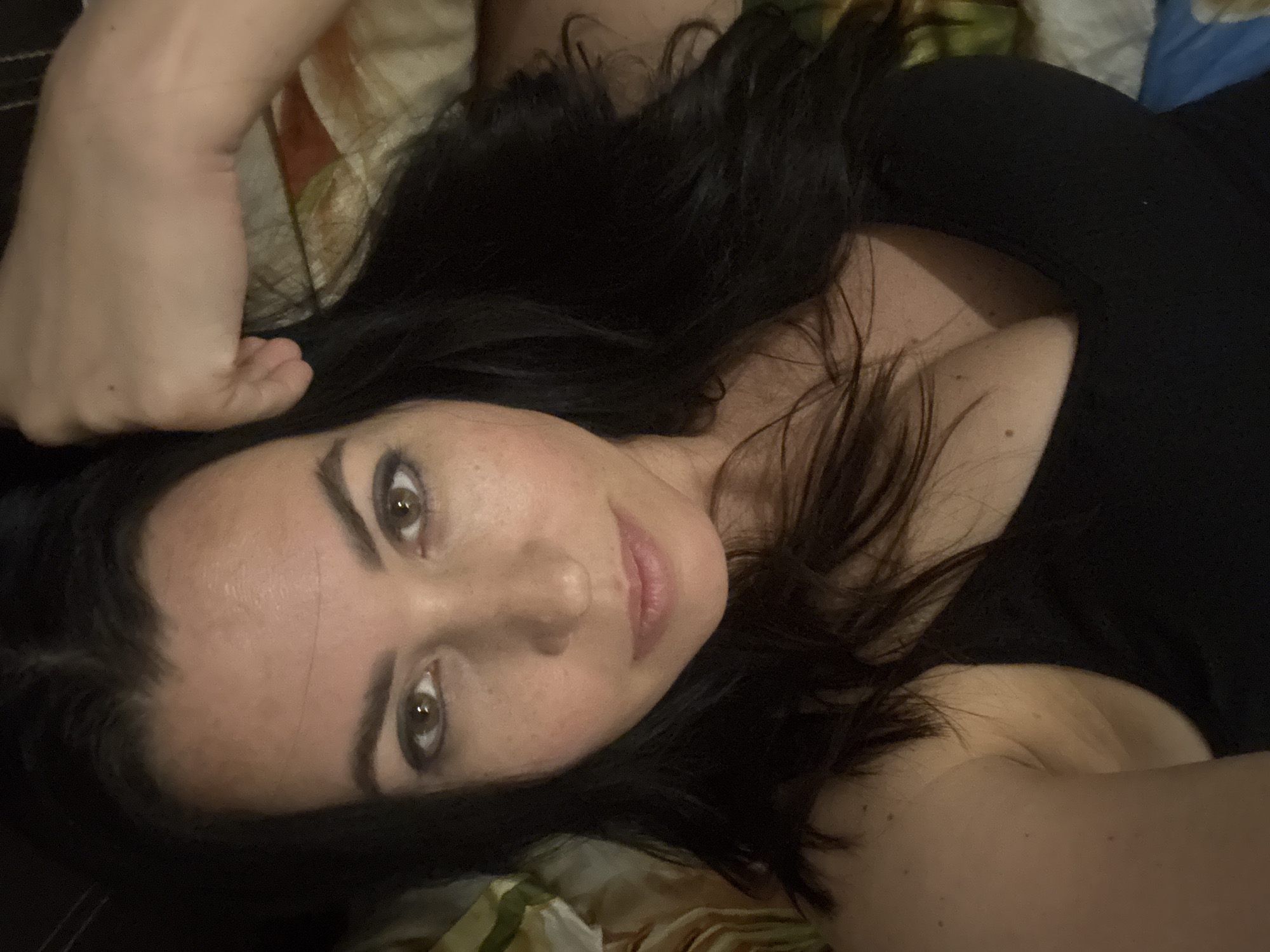 A woman with dark hair and light skin is lying down, facing the camera. She is wearing a black top. The background appears to be a bed with a patterned sheet. The location in Pattaya City, Thailand is not evident in the image.