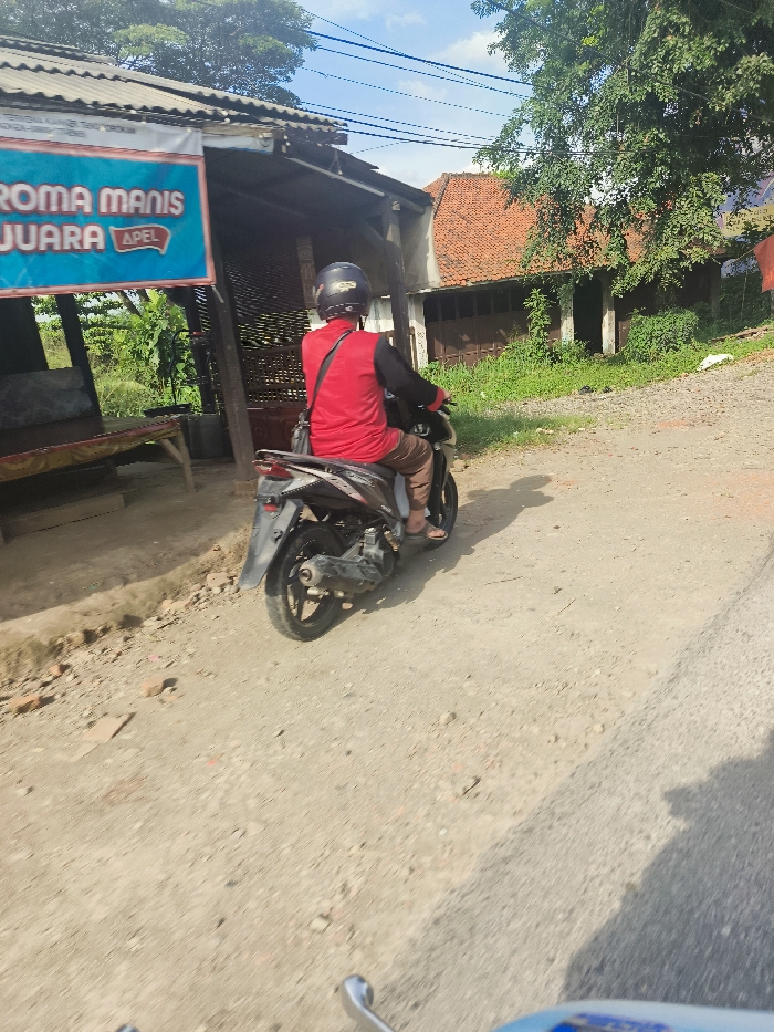 A man is riding a motorcycle down a dirt road in Jatiluhur, Indonesia. The man is wearing a red shirt and a helmet. He is riding on the right side of the road, heading towards the right. The motorcycle is a black Honda Beat. The road is unpaved and has a light dusting of gravel. There is a building to the left of the road. It has a brown wooden roof and a tan wall. The sign above the door says "Roma Manis Juara Apel". There is a small amount of grass and foliage to the left of the building. There is a house with a brown roof and a brown door on the right side of the image. The house is also in a state of disrepair. There is a lot of vegetation behind the house, with tall trees and a lot of shrubs. The sky is mostly clear with some white clouds. The location is -6.57 latitude, 107.438 longitude.