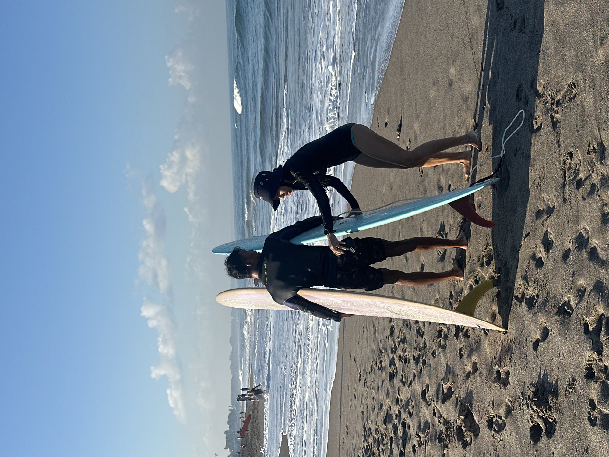 In North Kuta, Indonesia, a man and a woman stand on a sandy beach with surfboards. The man, wearing a black wetsuit and patterned shorts, holds a light-colored surfboard upright. The woman, in a black long-sleeved rashguard and blue bikini bottoms, wears a black bucket hat and grips a blue surfboard, which she is helping the man position. The surfboards are angled towards each other, creating a V-shape. The ocean stretches out behind them, with gentle waves and a white wash reaching the shore. The sky is a clear blue with a few scattered clouds. In the background, other people and surfboards are visible on the beach and in the water, suggesting a popular surfing spot. Footprints are imprinted in the dark sand.