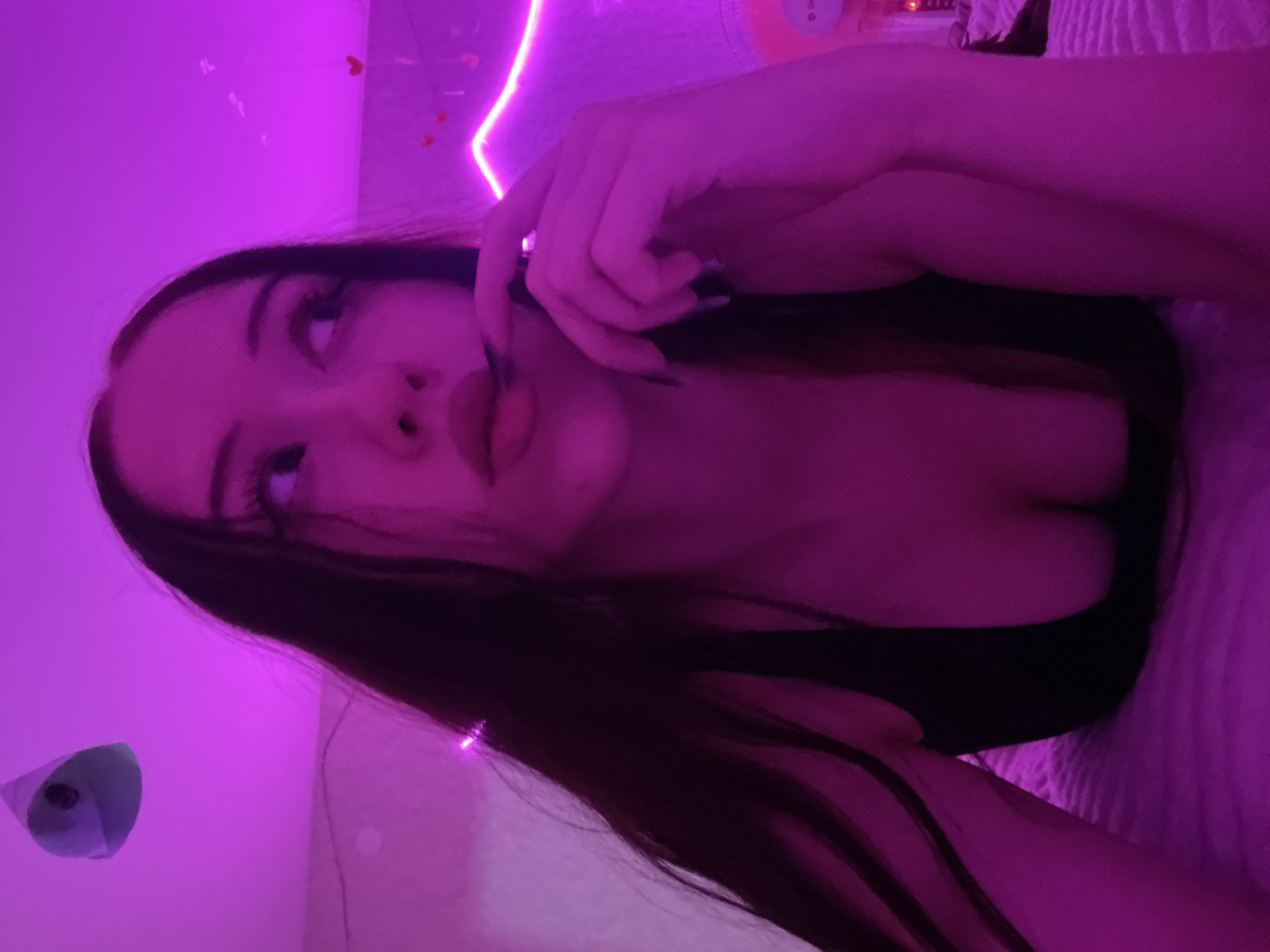 A young woman with long, dark hair is lying down in a room illuminated by pink and purple lighting. She is wearing a dark, low-cut top that reveals her cleavage. Her right hand is resting near her mouth, with her index finger touching her lips. The background of the room is decorated with pink neon lights in the shape of a heart and other small heart decorations. A rounded, light-colored object is visible hanging from the ceiling in the upper left corner of the image. The woman's expression is pensive as she looks to her right. The scene appears to be a bedroom or personal space.