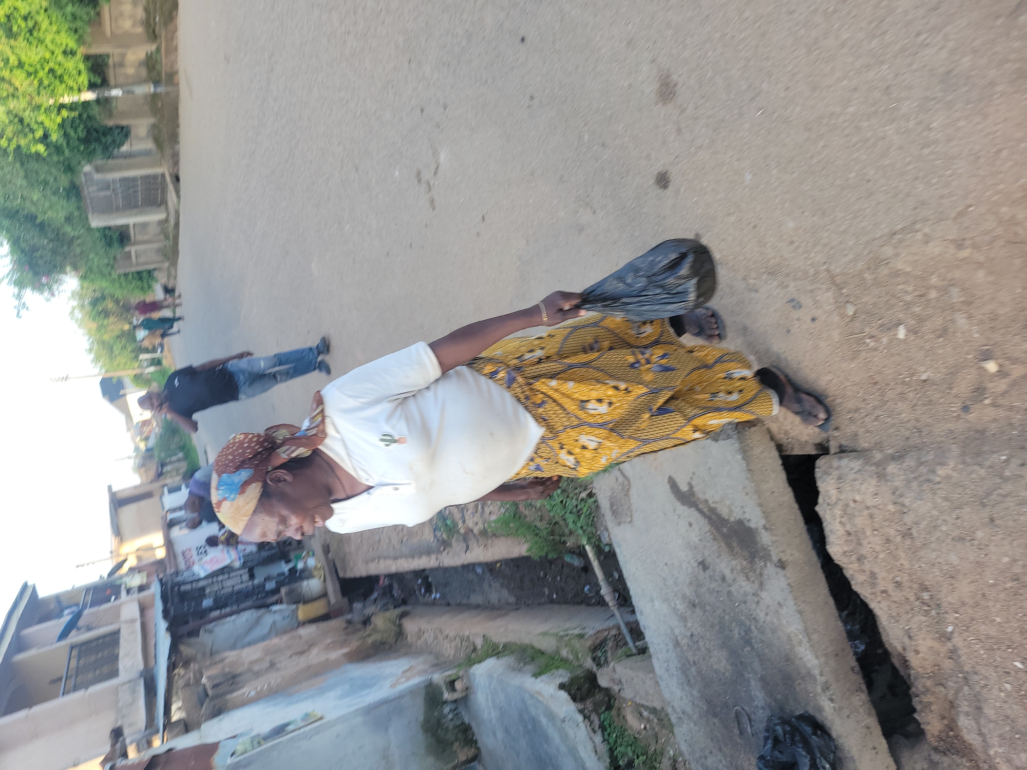 A medium shot captures a woman walking along a paved road in Jos, Nigeria. She is wearing a white polo shirt and a vibrant yellow and blue patterned skirt. A patterned headscarf adorns her head, and she carries a black plastic bag in her right hand. The woman is looking to her left, with her mouth open. To her left, a concrete drainage channel is visible, filled with murky water and debris. Behind her, a man in a blue shirt and jeans walks away from the camera, talking on a mobile phone. Further in the background, other pedestrians and buildings line the street, suggesting a residential or commercial area. The overall lighting indicates it is daytime, with natural light illuminating the scene. The environment appears somewhat unkempt, with visible dirt and some litter.