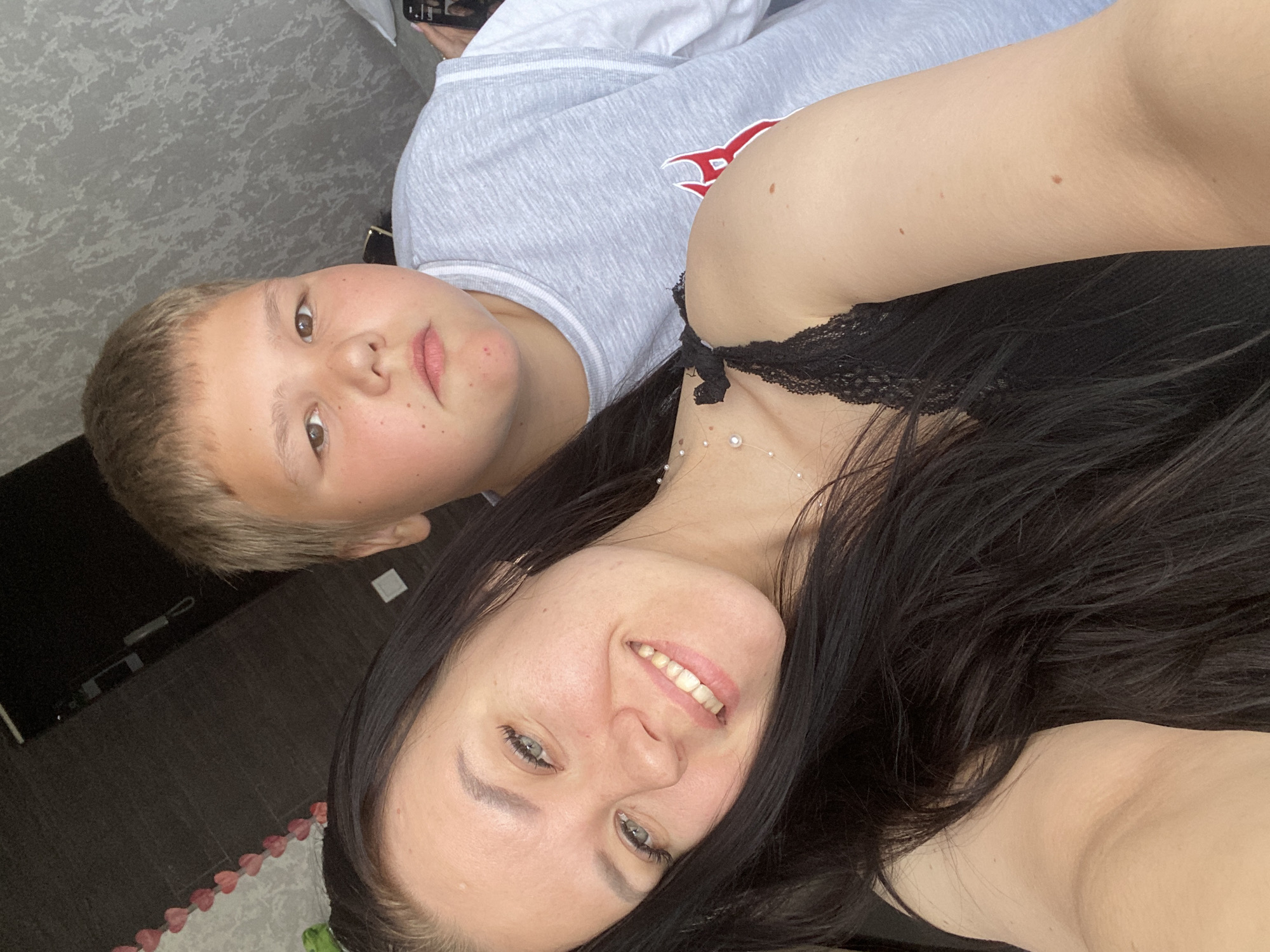 A woman with long, dark hair, wearing a black top with lace trim, and a young boy with blonde hair and freckles, wearing a gray sweatshirt with a red emblem, are posing for a selfie. The photo appears to have been taken indoors. A TV can be seen on the wall. The location cannot be confirmed as the city of Chaikovsky, Russia based on the image.