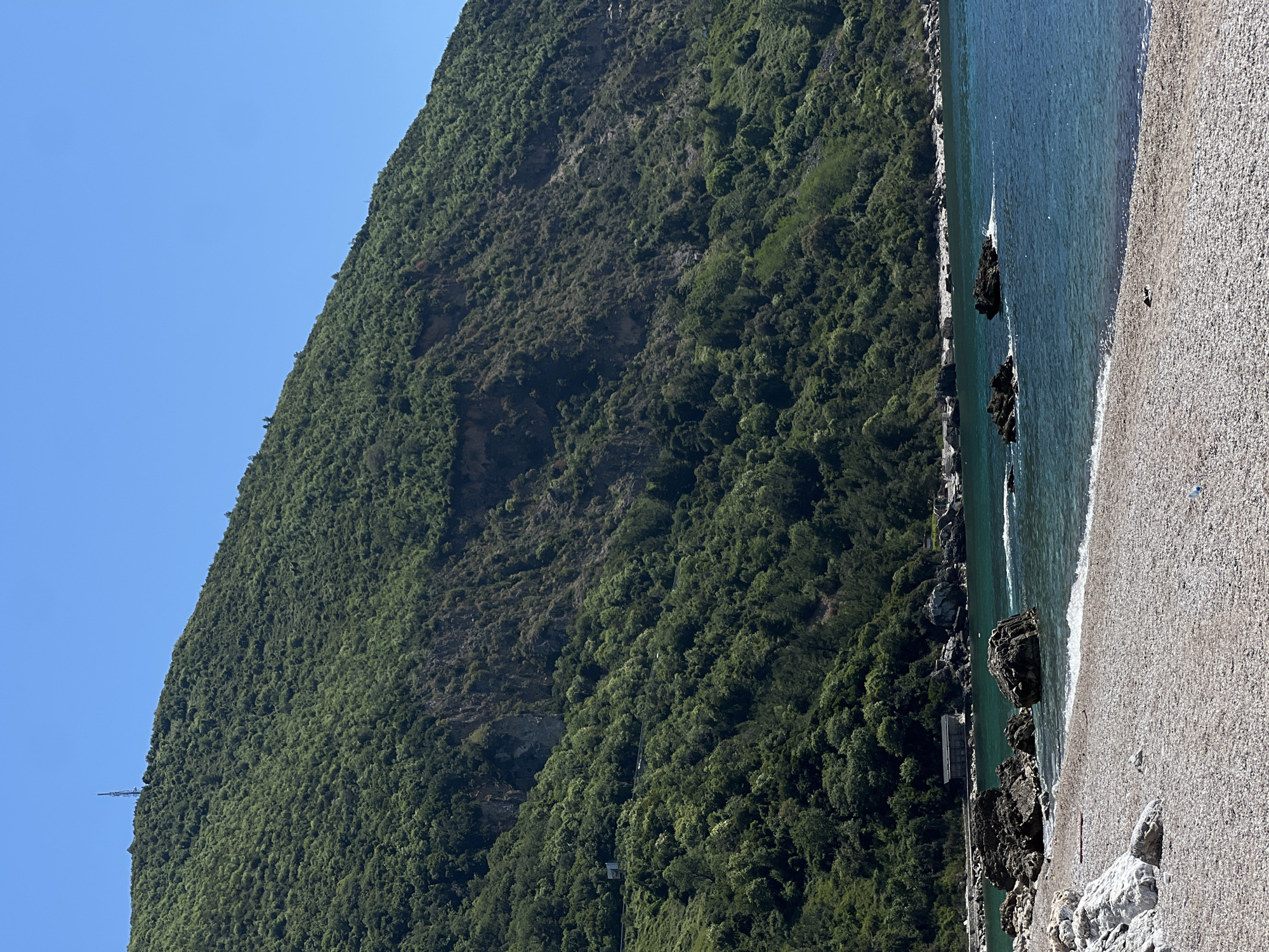 The image depicts a coastline in Prijevor, Montenegro, with a steep, densely forested mountain on the left side. A gravel beach runs along the right side, meeting clear turquoise water with scattered dark rocks. A utility pole or tower can be seen at the top left of the mountain.