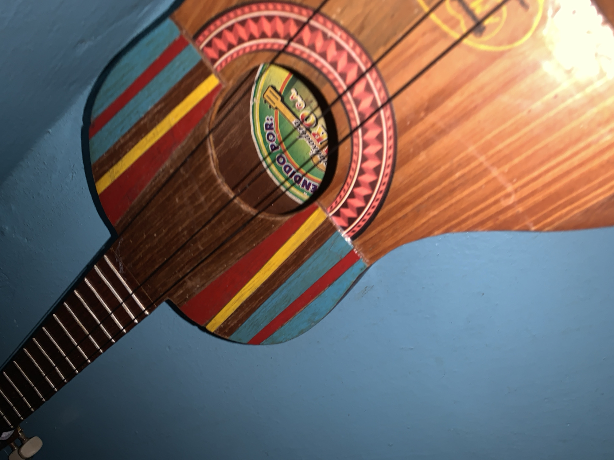 The image is a close-up, high-angle shot of a colorful, wooden string instrument resembling a guitar, leaning against a textured, light blue wall. The instrument features a brown wooden neck with white frets and tuning pegs visible at the top. The body of the instrument is adorned with vibrant, diagonal stripes of red, yellow, and blue. A circular soundhole is present on the body, featuring a colorful decal with text that appears to read "VENDIDO POR:" and "KRAUSSA". The surrounding area of the soundhole has a decorative inlay of red and white geometric patterns. The wood of the instrument shows a visible grain pattern and has a glossy finish. The blue wall provides a plain background, with some subtle shadowing suggesting an indoor environment. The scene is static, with no people or overt actions depicted. This scene is not specifically located in Santa Ana, Venezuela, as no geographic markers are present within the image.