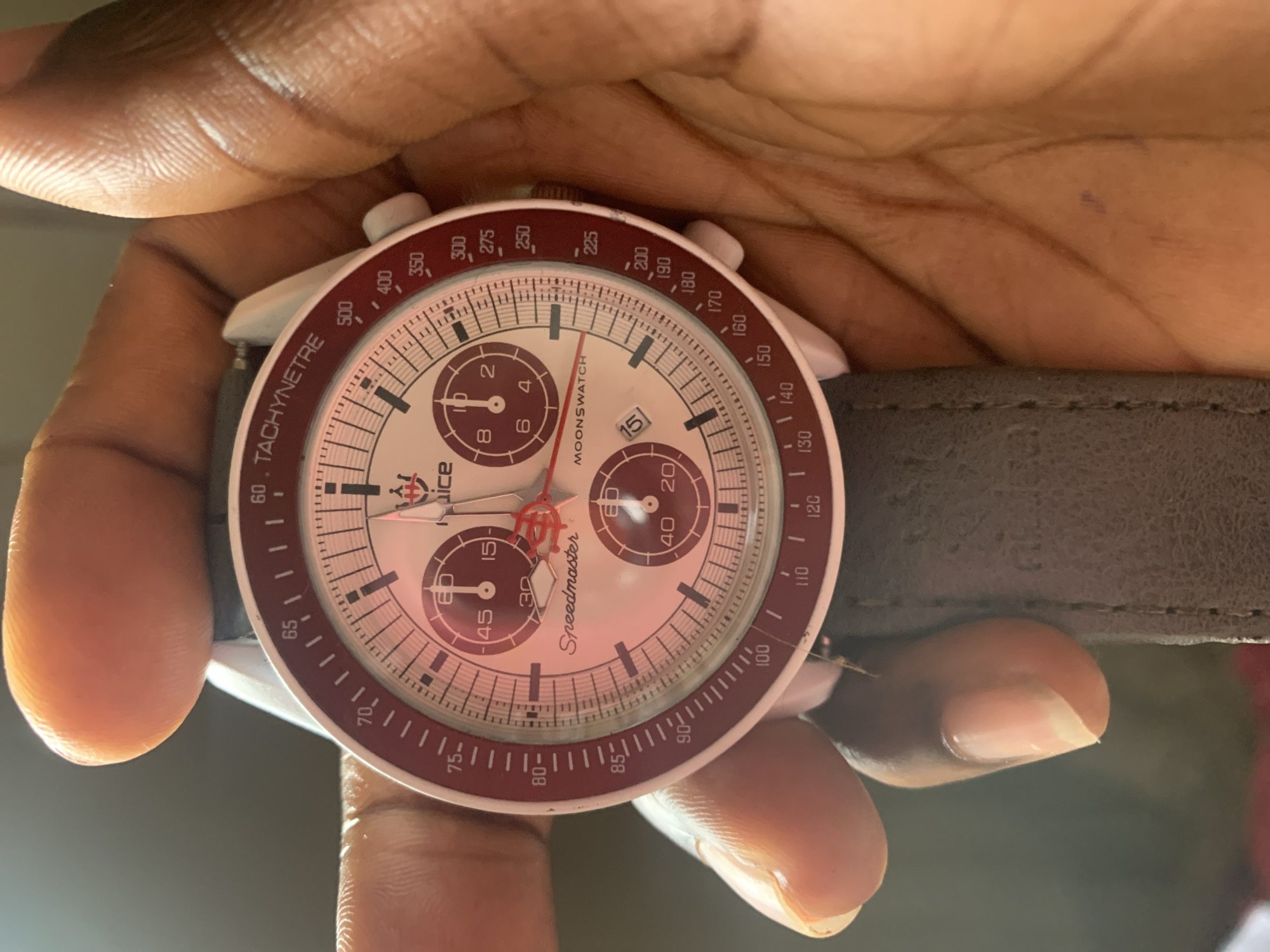 A person's hands are holding a white and red watch with a dark gray strap. The watch has a white face with red accents and sub-dials. The words "Speedmaster" and "MOONSWATCH" are visible on the face. The outer bezel of the watch is red and has tachymetre markings. The background is dark and out of focus, suggesting an indoor setting. There are no specific landmarks or context to determine a city or location.