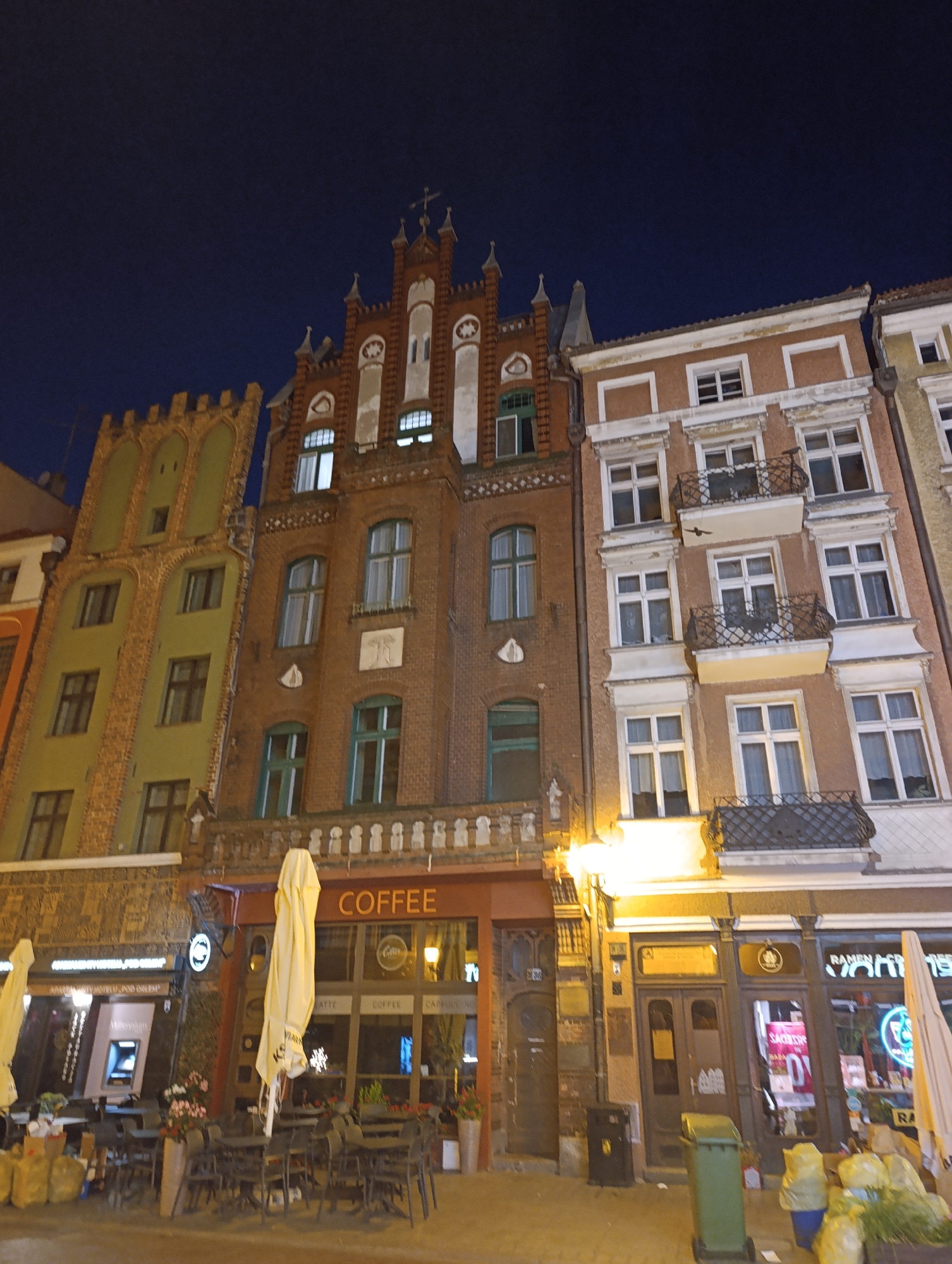 The image depicts a street scene in Toruń, Poland, at night. In the foreground, outdoor seating areas for cafes and restaurants are visible, with tables and chairs set up. Several large umbrellas are present, some open and some closed. Various items, including bags of trash and potted plants, are scattered around the seating areas.
The buildings lining the street are multi-story structures with distinct architectural styles. On the left, a pale green building with arched window frames stands next to a red brick building with a more elaborate facade, featuring decorative white elements and a cross at its peak. Further to the right, a peach-colored building with white trim and balconies is visible. One of the businesses on the ground floor has a prominent "COFFEE" sign. Another establishment to the left displays signs and what appears to be an ATM. The street is illuminated by artificial lights, casting a warm glow on the scene. The sky above is dark, indicating it is nighttime.
