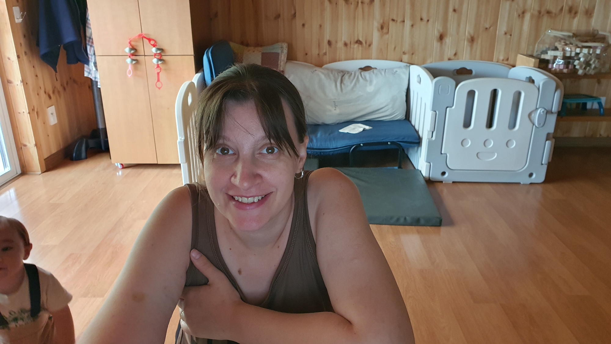 In an indoor setting, a woman with medium-length brown hair and bangs smiles directly at the camera. She is wearing a brown tank top and has hoop earrings. Her right arm is crossed over her chest. To the left of the woman, a young child wearing overalls is partially visible. The background features a wooden wall and floor. There is a light-colored cabinet with some decorative red items hanging on it. A white and gray playpen with a smiley face design is positioned in the middle of the frame. Behind the playpen, a blue mat and a pillow are visible. A shelf unit with various items is located to the right of the playpen. The scene appears to be a living area or playroom.