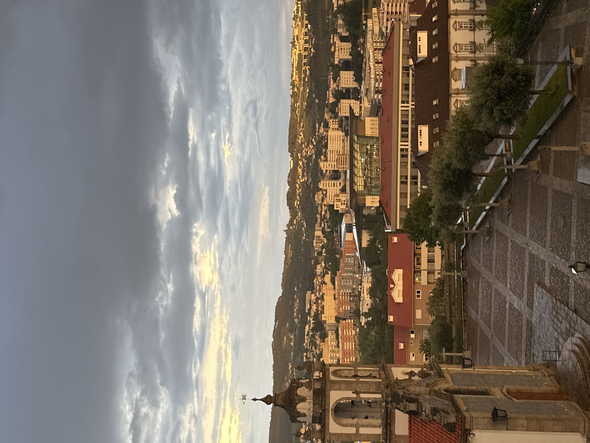 This image captures a panoramic view of Vila Real, Portugal, at what appears to be late afternoon or early evening, characterized by dramatic lighting from a setting or rising sun. The scene overlooks an expansive cityscape built into undulating hills, under a sky with a mix of heavy, dark clouds and brighter, illuminated patches.
In the foreground, a prominent stone church tower with ornate architectural details, including a spire topped with a weather vane, anchors the right side of the frame. Adjacent to the church is a large, geometrically patterned cobbled square, which leads into linear planted areas featuring evenly spaced, well-maintained trees. The architecture of the buildings immediately surrounding the square includes traditional structures with distinctive windows and roofs.
The mid-ground and background showcase a dense urban sprawl, with numerous buildings extending across and up the hillsides. Many of these structures, ranging from multi-story residential blocks to more modern commercial buildings, are bathed in a warm, golden light, creating a striking contrast with the darker areas. Red-tiled roofs are a common feature among the buildings, interspersed with areas of greenery and trees. The distant hills are covered with a blend of natural vegetation and urban development.
The sky transitions from a deep, somber grey on the left, indicative of heavy cloud cover, to lighter, more ethereal clouds on the right, where the sun's golden rays are breaking through. This dynamic sky contributes significantly to the atmospheric quality of the image. The overall impression is one of a bustling city nestled within a natural landscape, captured during a serene and visually striking time of day. No visible text is present in the image.