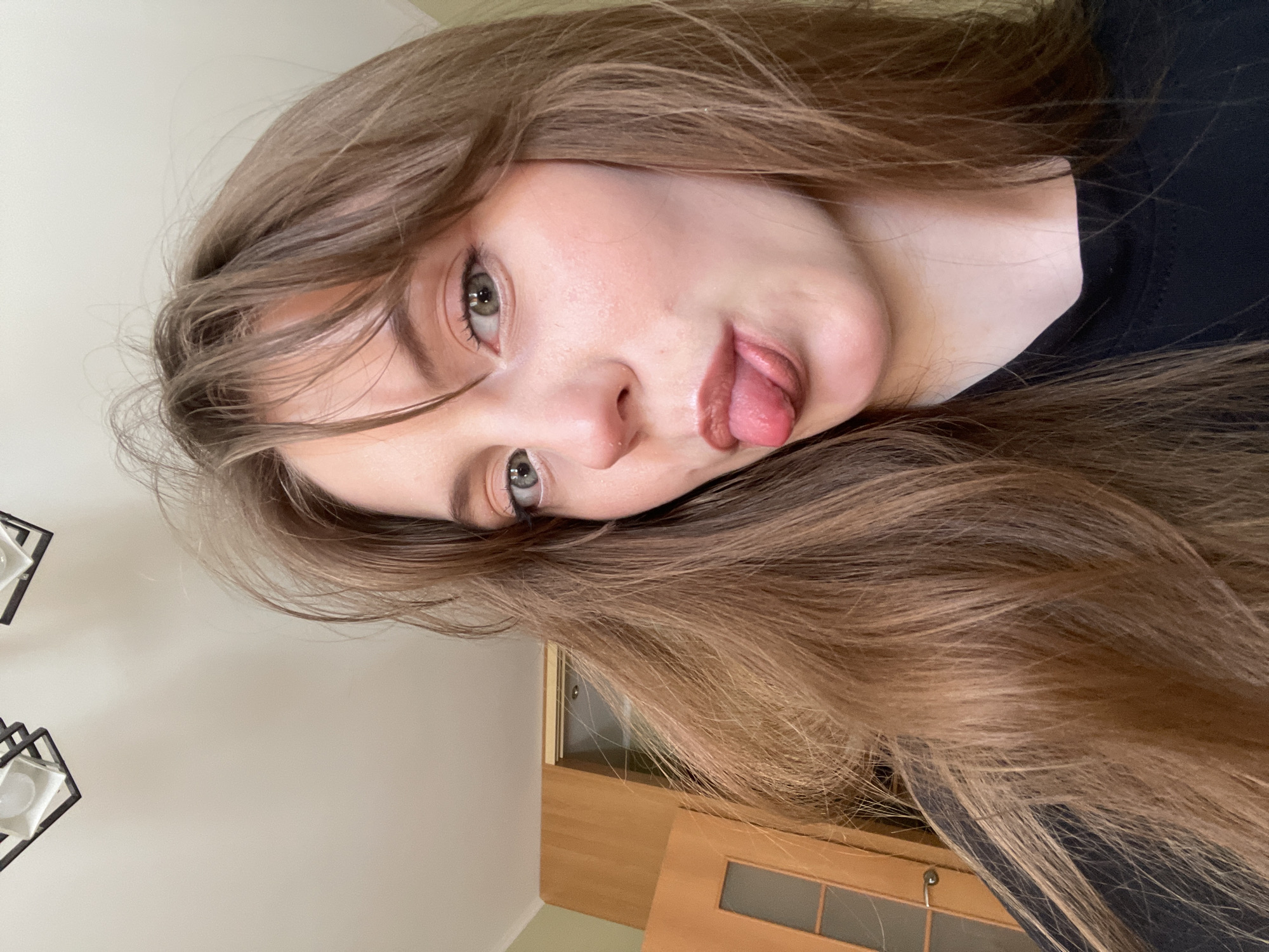 A young woman with long, light brown hair and fair skin is centered in the frame. She is wearing a black shirt. Her face is angled to the left, and she is sticking her tongue out playfully. Her eyes are open, and she is looking at the camera.
The background reveals part of an interior. A beige wall is visible in the top left corner, while a light brown wooden cabinet or door with glass panels occupies the lower right. A modern black light fixture is partially visible in the top left.
There is no discernible information to confirm the location being in Ufa, Russia.