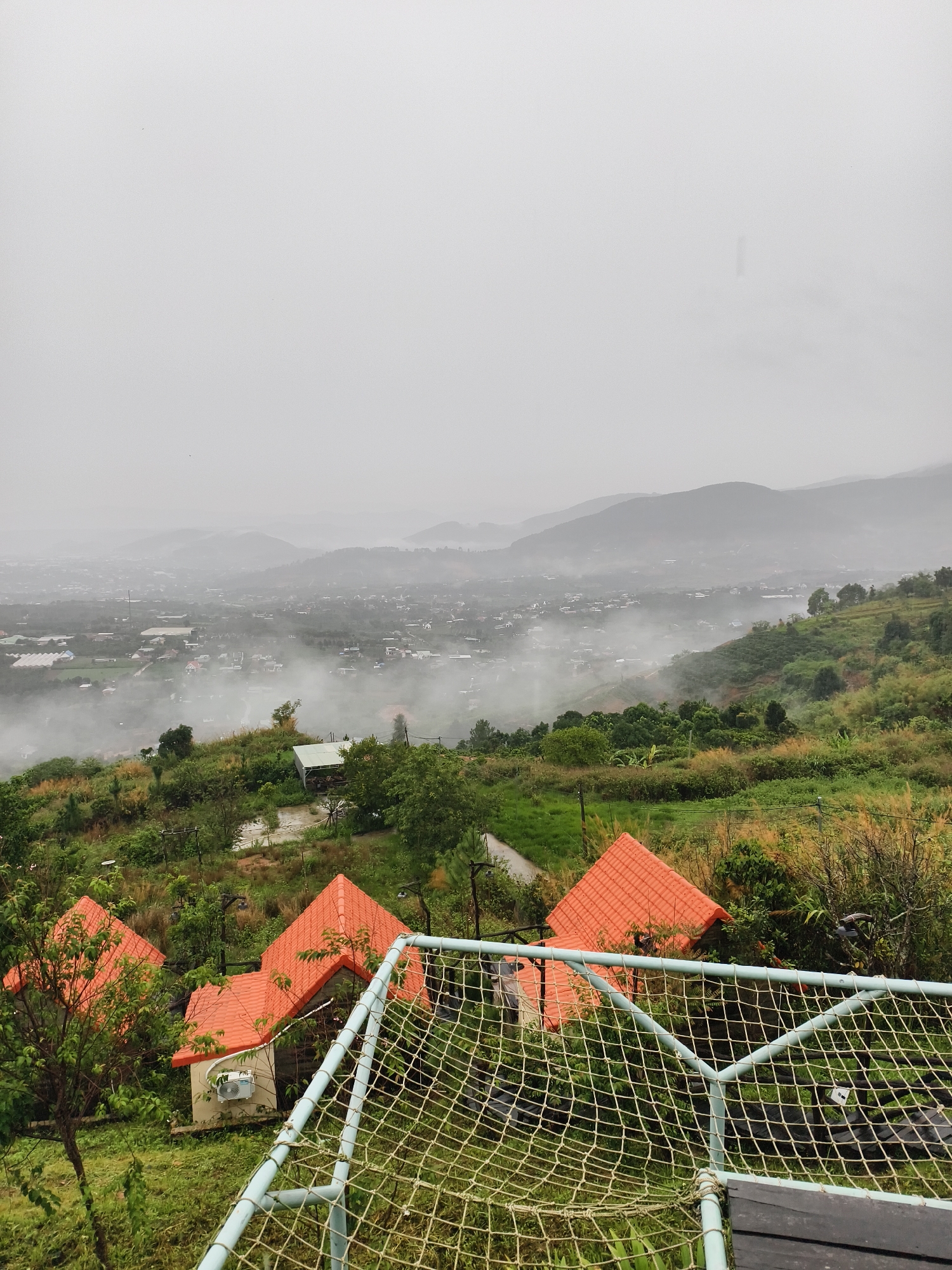 The image presents a high-angle view of a landscape shrouded in mist, with buildings featuring orange roofs scattered across a green, hilly terrain. A safety net structure is visible in the foreground, and mountains fade into the hazy background under an overcast sky. The location appears to be rural or suburban, blending natural and built environments. Specific details like precise locations and identifying features are obscured by the fog.