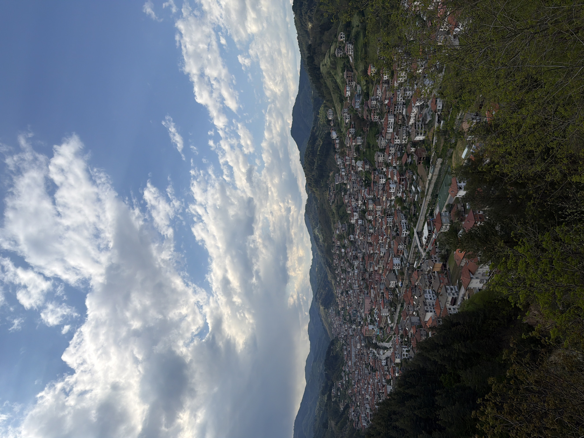 The image depicts an overview of a densely populated town in Nedelino, Bulgaria, situated on a hillside. The town consists of numerous buildings with reddish-brown roofs. The sky above shows a mix of blue and clouds. Green foliage and trees surround the edges of the town, indicating a natural environment. In the background are mountains.