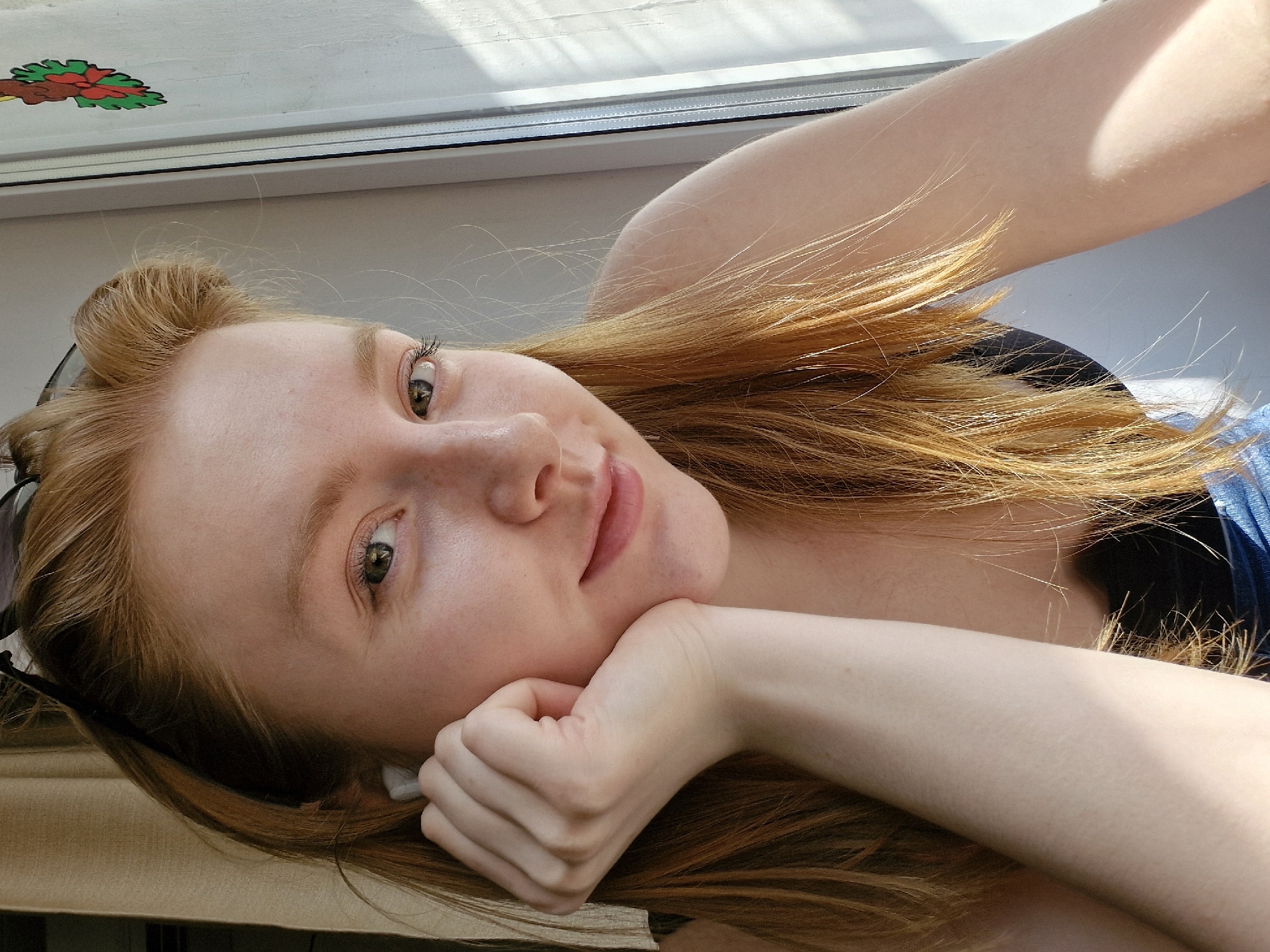 A young, fair-skinned woman with red hair is lying down indoors, propped up on her right hand. She is looking at the camera with a slight smile. Her hair is spread out around her head and arm. She is wearing a dark-colored top with a glimpse of blue fabric visible. She is positioned near a window frame with a sticker in the upper left corner. The scene is brightly lit, likely from the window. The location cannot be determined to be Yekaterinburg, Russia based on the image.