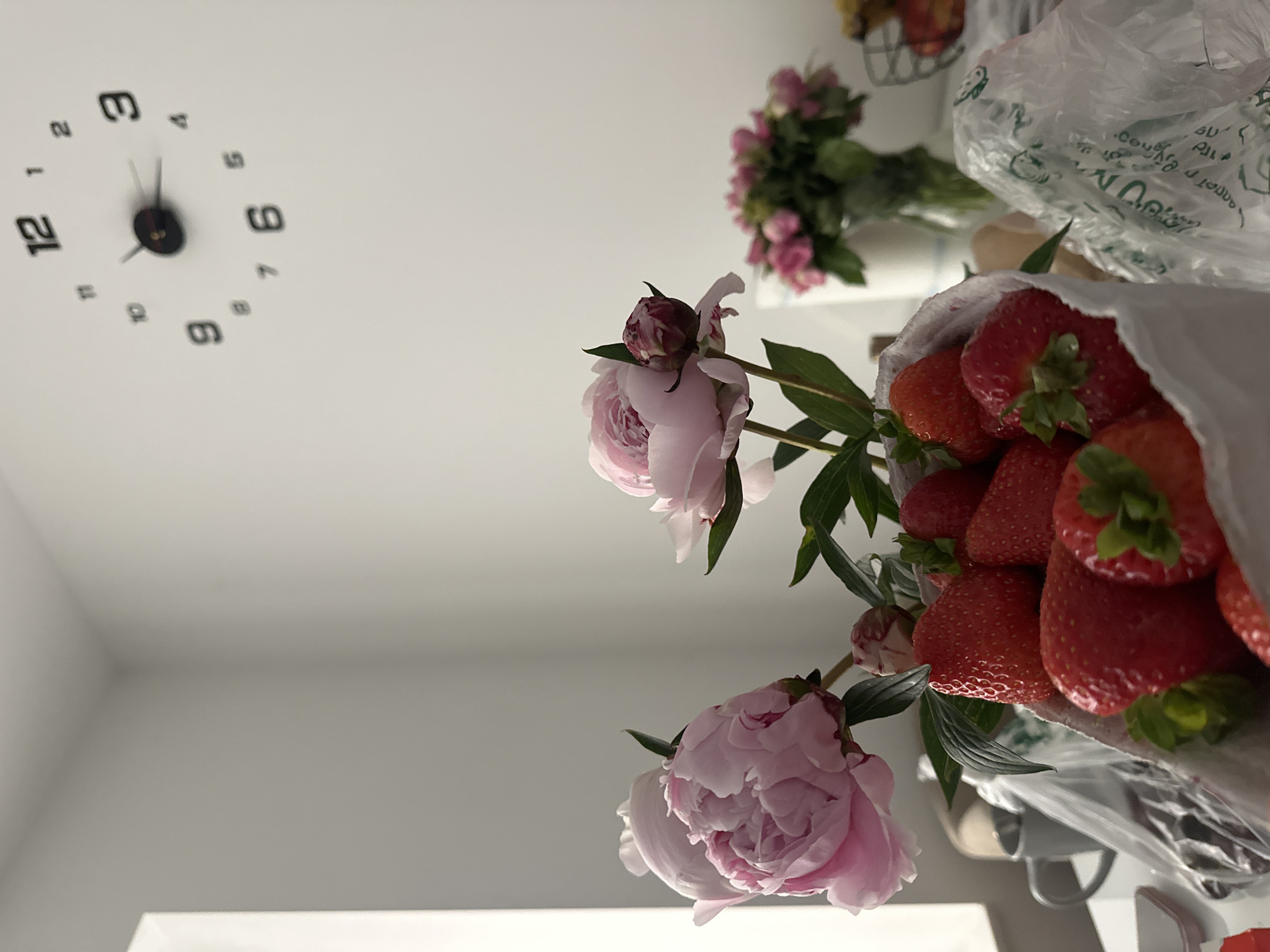 The image depicts a still life arrangement on a table, featuring a cluster of fresh strawberries in a white paper bag and two large pink peonies. In the background, a modern wall clock with black numerals is visible on a white wall. A vase with a bouquet of pink flowers and a metal fruit basket containing apples are also present. The scene appears to be indoors, possibly in a home environment in Kutenholz, Germany, given the context provided. There are no people visible in the image, and no actions or interactions are taking place. The overall lighting suggests it is daytime.