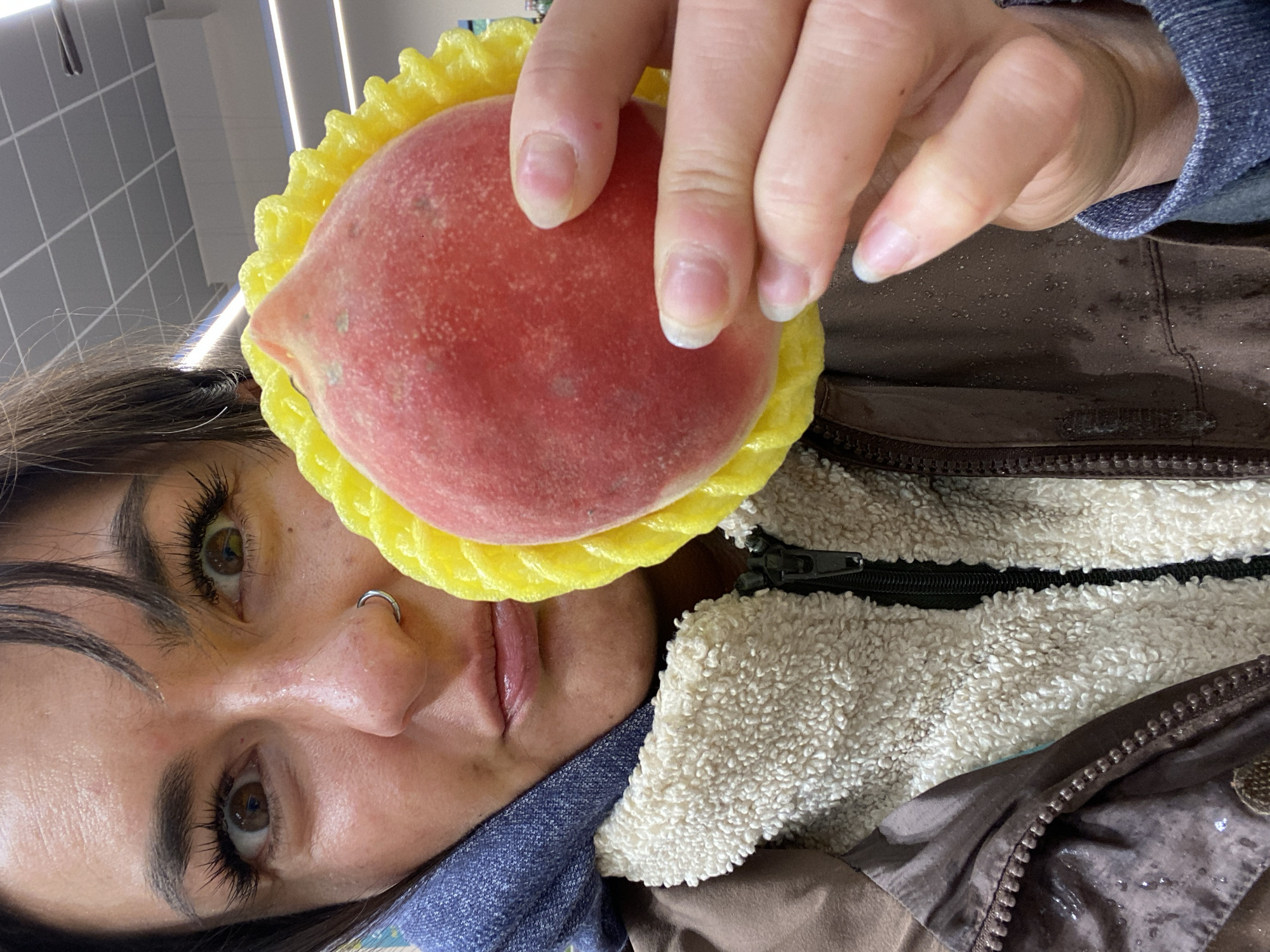 A woman with light brown eyes and dark hair is holding a red peach with a yellow foam protector in front of her face. She has a silver nose ring and long eyelashes. She is wearing a brown jacket with a white fuzzy interior and a blue shirt underneath. The background is out of focus and shows a tiled wall and a light fixture. There is no information available to determine if this image was taken in Moscow, Russia.