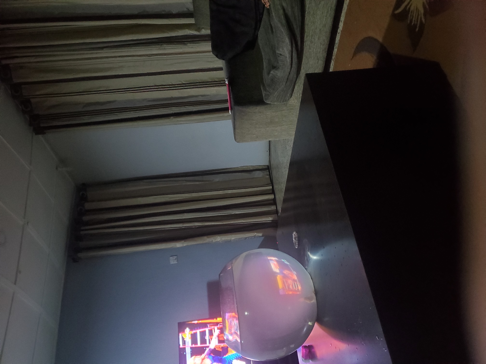 This image depicts an indoor scene, likely a living room, with a dark wooden coffee table in the foreground. On the table, a clear, round fishbowl filled with water is prominently featured. To the left of the fishbowl, a small pink object is visible. In the background, a television screen displays an image of a woman with a ladder. To the right of the television, a light blue wall is present, with a light switch on it. Further to the right, beige curtains with dark vertical stripes hang from a curtain rod. A portion of a couch, upholstered in a greyish-brown fabric, is visible on the far right, with a dark cushion on it. A red object, possibly a lighter or pen, rests on the armrest of the couch. The floor appears to be tiled, with a patterned rug in the lower right corner. The lighting in the room is dim, creating shadows and reflections. The image is taken from a slightly low angle, looking towards the right. No specific details about the city of Jos, Nigeria, are visible within the image itself.