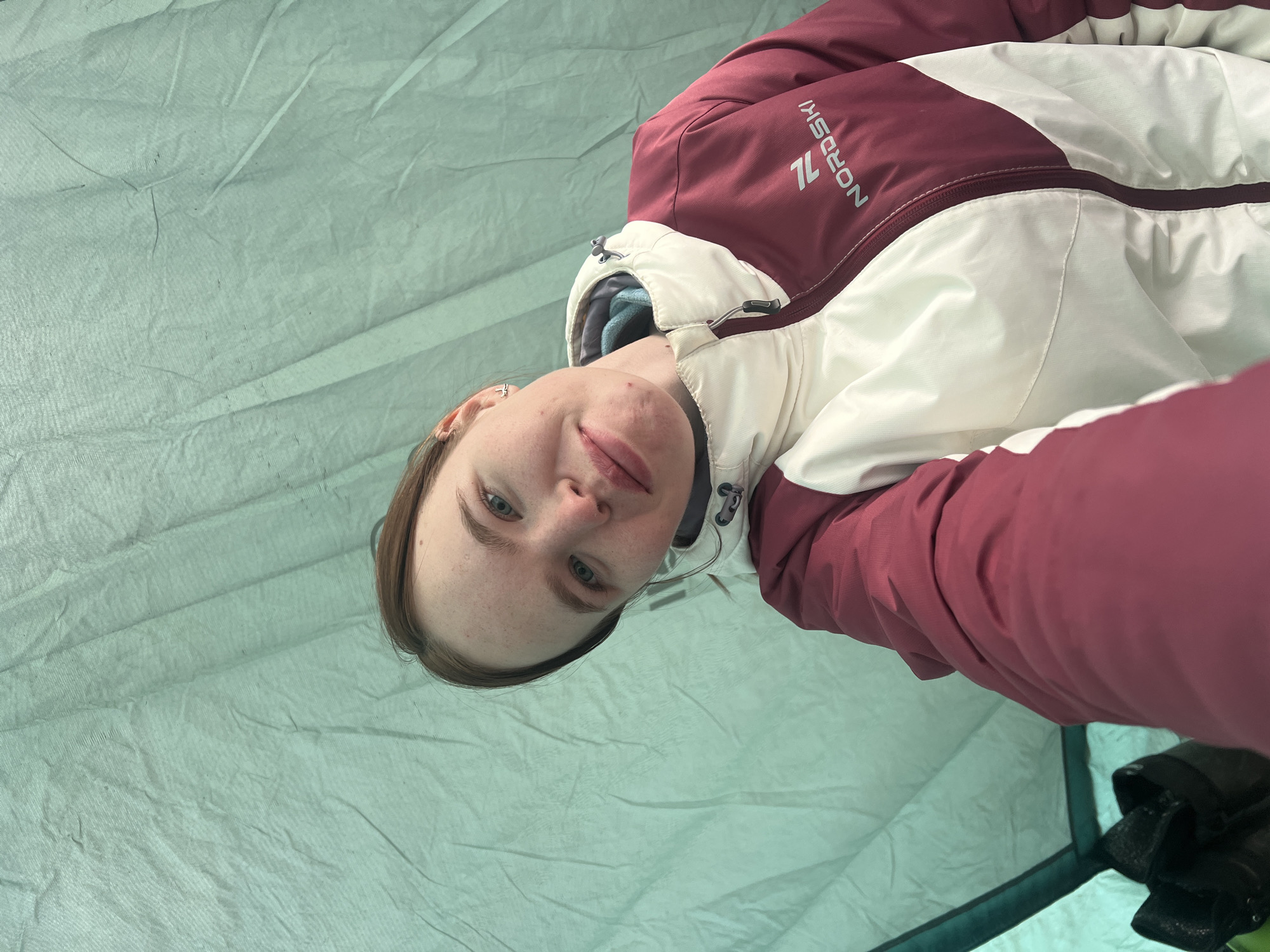 A close-up selfie shows a young woman lying down inside a tent. She has fair skin, light blue eyes, and light brown hair, with visible freckles across her face. Her expression is neutral and calm, possibly with a slight, subtle smile. She wears a warm, insulated jacket that is predominantly maroon/dark red and off-white/cream, with a visible light blue inner collar. The words "NORDSKI" and the number "7L" are visible in white lettering on the maroon section of her jacket. A small silver earring is visible in her left ear, and there is a faint reddish mark near her left eye and on her lower lip. The interior of the tent forms the background, made of a light green or teal fabric that shows creases and a slightly textured, possibly mesh-like, appearance. In the bottom right corner, a dark green tent edge and some black objects, likely shoes or bags, are partially visible. The scene suggests an outdoor activity such as camping in Lesnoye, Russia.