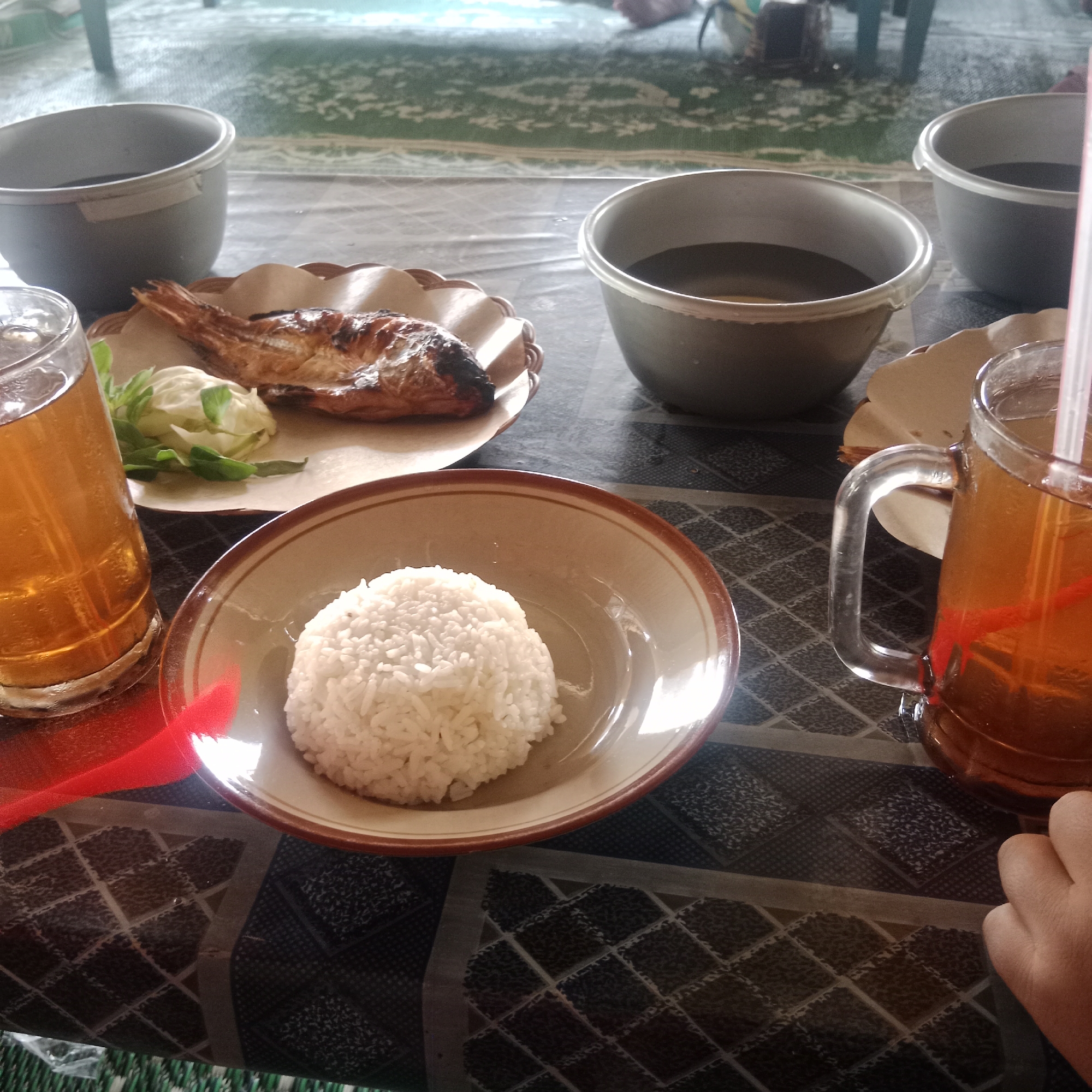 This is a close-up, eye-level shot of a meal set on a patterned tablecloth. In the foreground, a glass of iced tea with a straw sits to the right of a bowl of white rice. To the left of the rice bowl is a plate with a grilled fish and some lettuce. In the background, two grey bowls containing dark liquid are visible, along with a blurred green and white patterned carpet. The setting appears to be a casual dining area, possibly outdoors given the lighting. The image does not contain any specific elements that place it in Ngandul, Indonesia.