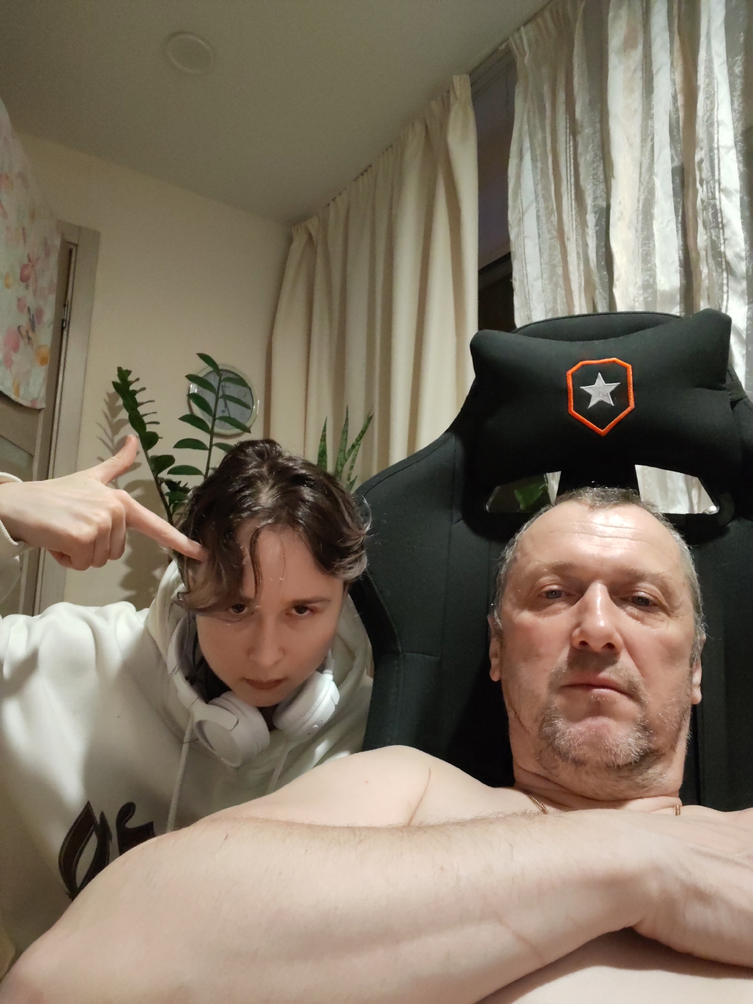 Two people are in a room with light-colored walls and curtains. A younger person in a white hoodie and headphones is pointing at their head. An older, shirtless person is sitting in a black gaming chair with an orange star logo. There are plants in the background. The scene appears to be indoors and casually posed. The location is not discernible from the image.
