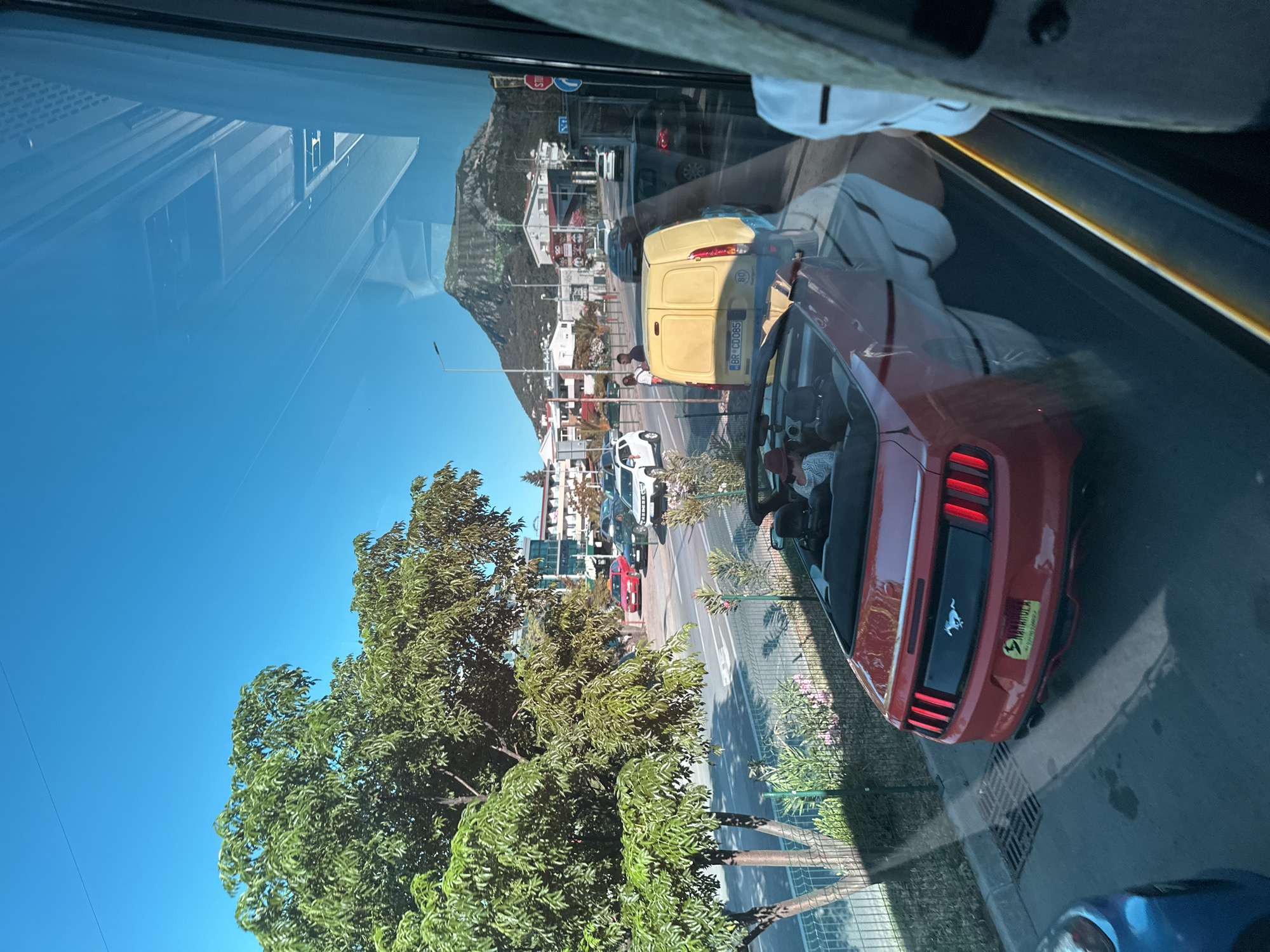 The image shows a street scene in Sutomore, Montenegro, viewed through a reflective window, likely from a vehicle. In the foreground, a red convertible, possibly a Ford Mustang, is parked. A person wearing a hat is seated in the driver's seat. The street is lined with trees, bushes, and a fence. Several cars are visible on the road, including a white SUV, a red sedan, and a yellow van. In the background, buildings and a large, green mountain are visible under a clear blue sky. The reflections in the window show parts of the interior of the vehicle from which the photo was taken, including what appears to be a person's arm and torso.