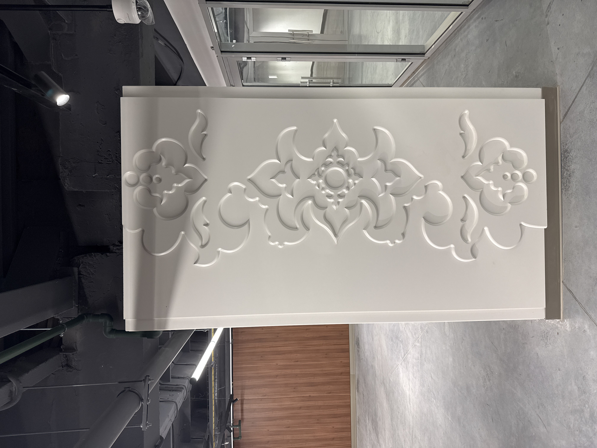 The image shows a white panel with a floral relief design in an interior setting. The panel is positioned below a window and adjacent to what appears to be a wooden door or wall panel. Above the panel, there are pipes and a surveillance camera mounted on a dark grey ceiling. The floor is a light grey, textured surface, likely concrete. The location is inferred to be Kazan, Russia, based on the context provided.