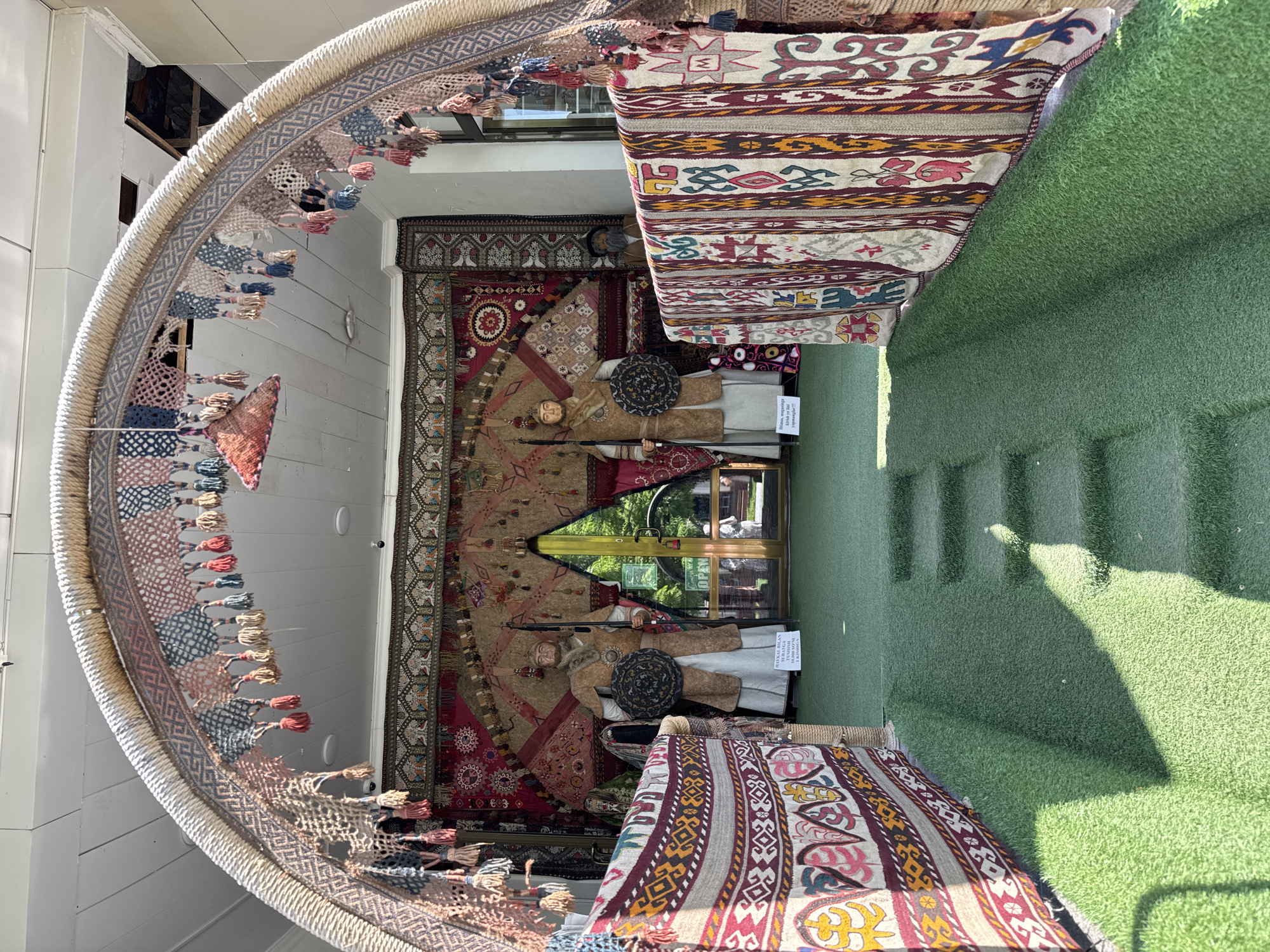 The image shows an interior setting in Samarkand City, Uzbekistan, displaying cultural artifacts. Mannequins dressed in traditional clothing are placed against a backdrop of ornately patterned rugs and carpets. Above, a semi-circular structure adorned with tassels and woven patterns hangs, creating a decorative element. The space includes what appears to be artificial green turf on the ground and possibly a building entrance, visible through a glass door or window. The scene aims to showcase the cultural heritage of the region through its textiles and traditional garments.