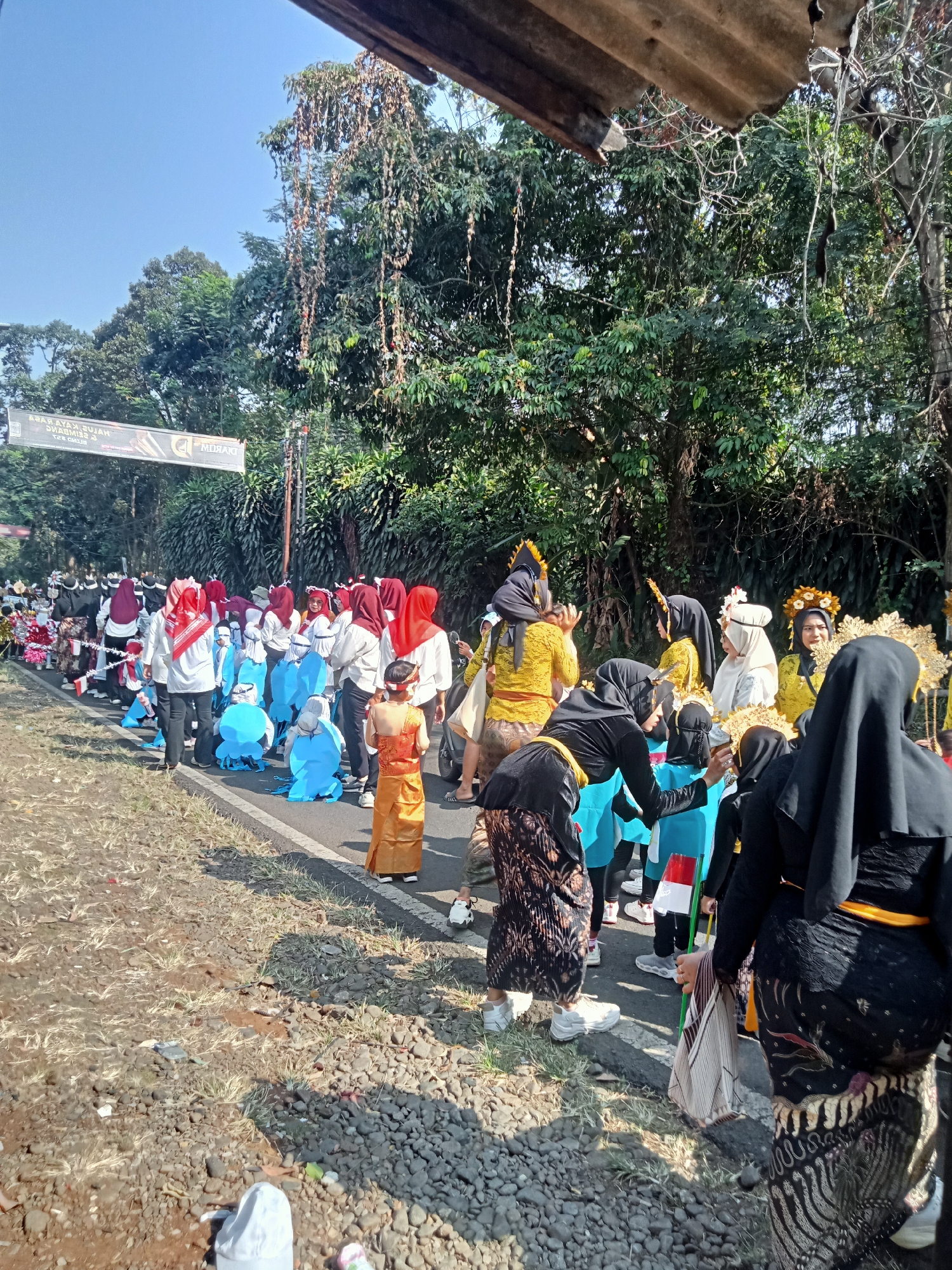 In Dororejo, Indonesia, a procession of people in colorful traditional attire walks along a roadside. The individuals are dressed in a variety of outfits, including brightly colored robes, headscarves, and elaborate headwear adorned with gold accents. Some participants wear blue costumes, possibly representing a specific theme or group. A banner with Indonesian text is visible in the background, suggesting a festive or ceremonial event. The road is lined with trees and vegetation, indicating an outdoor setting in a semi-urban or rural area. The ground is covered with gravel and dry grass, with a paved road forming the path for the procession. The sun is shining, casting shadows and illuminating the scene.