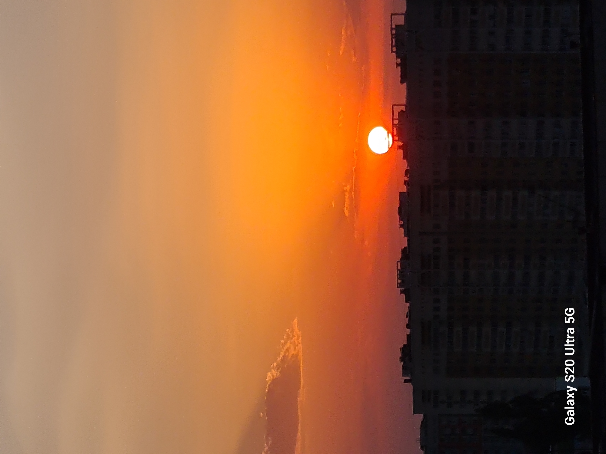 The image captures a sunset over the city of Almaty, Kazakhstan. The sun, a bright white orb with an orange glow, is partially obscured by the rooftops of silhouetted buildings in the foreground. The sky displays a gradient of warm colors, transitioning from a soft peachy hue at the top to a vibrant orange and then a deep red near the horizon. A few wispy clouds are visible in the sky, catching the sunlight. The buildings appear to be apartment blocks, their dark silhouettes stark against the colorful sky. The lower right corner of the image features the text "Galaxy S20 Ultra 5G", indicating the device used to capture the photograph.