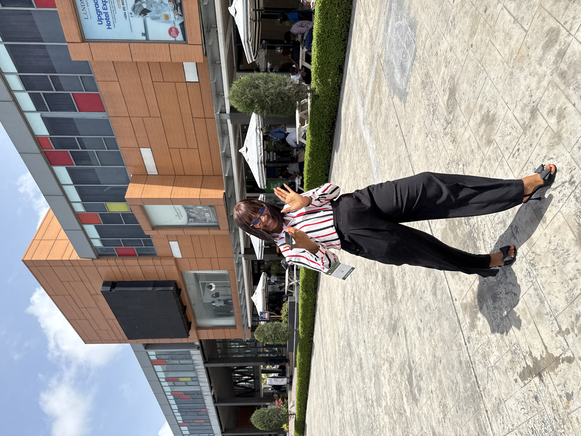 Here's a forensic description of the image:
**Scene:** The image captures a woman standing in a paved public space. Behind her is a modern building, identifiable by its architectural style and signage, indicating the location is in Alaguntan, Nigeria.
**People:** A single female is centered. She is wearing sunglasses, a horizontally striped shirt, and black wide-leg pants. She has short brown hair. Her right hand is raised near her face, palm outwards, in a waving gesture.
**Objects:** The environment contains architectural details, including windows, awnings, and signage for a hotel. There is signage visible indicating "HOMERE" and "EXHIBITOR" on what appears to be a lanyard. The woman is wearing sandals with bow details.
**Actions:** The woman is posing for the photo, raising her hand in greeting.
**Environment:** The scene appears to be in an outdoor plaza. Tables and chairs are present behind the woman, suggesting a dining area or public gathering space. The weather appears sunny based on the shadows.
