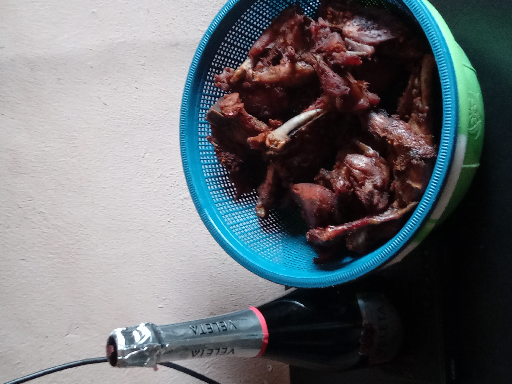 The image depicts a blue plastic basket filled with dark, fried poultry pieces, including bones. The basket sits near a bottle of "VELETA" labelled sparkling wine. The background is a light-colored wall on the left and a dark surface on the right. It is unclear if this is in Sabon Tasha, Nigeria.