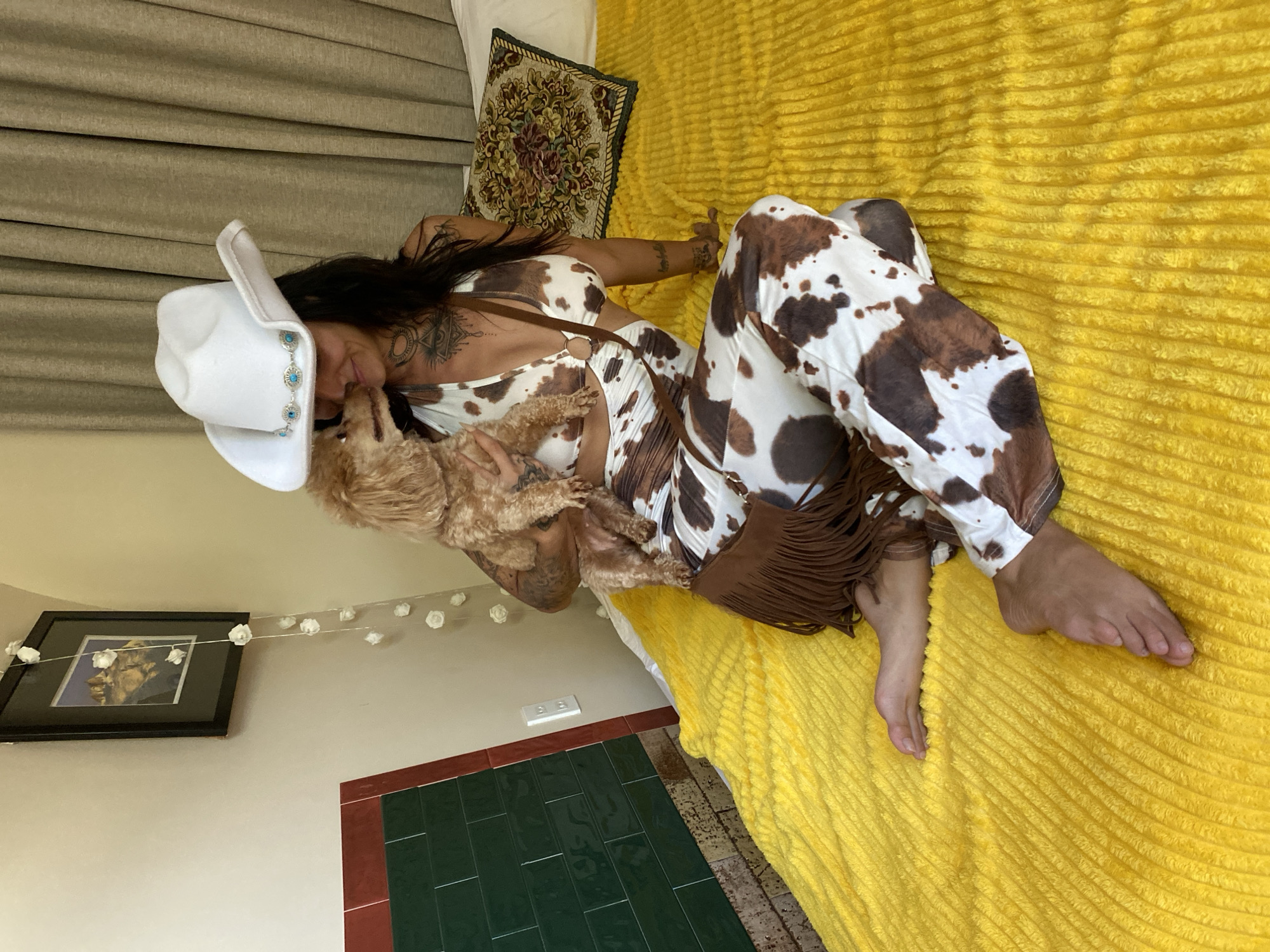 A woman wearing a white cowboy hat and a cow-print outfit sits on a bed covered with a yellow, textured blanket. She is holding a small, light brown dog. A brown fringed bag hangs from her shoulder. To the left of the bed, there is a framed picture on the wall and a section of green tiled wall with a red trim. Behind the woman, a curtain is visible. The setting appears to be an indoor location, possibly a bedroom, and there is no indication of it being in Mũi Né, Vietnam.