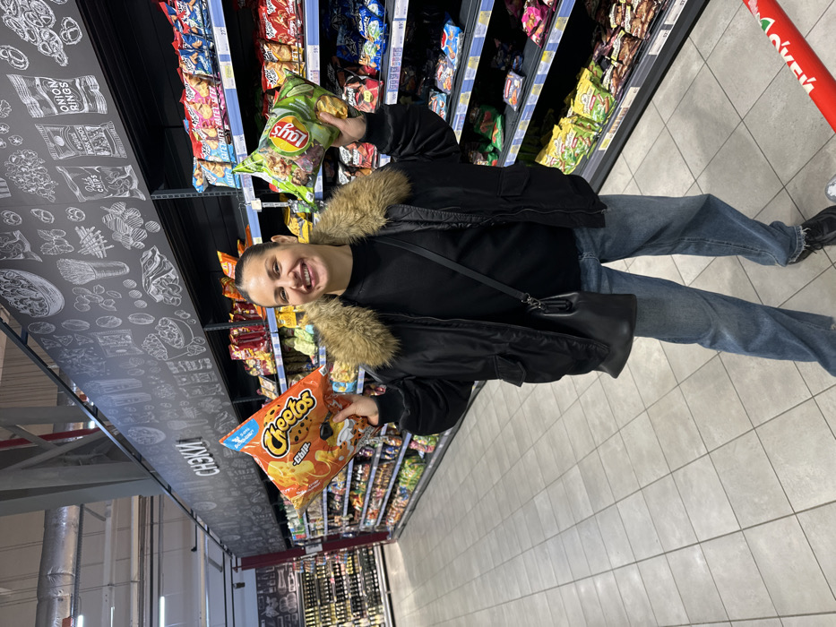 An adult female stands in a supermarket aisle in Sochi, Russia. She is facing the camera, smiling broadly. The subject wears a black winter jacket with a brown fur-lined hood, a black long-sleeved top, light blue denim jeans, and dark athletic shoes. A black shoulder bag with a thin strap is slung across her body. She holds a large orange bag of Cheetos in her left hand and a large green bag of Lay's potato chips in her right hand, presenting them towards the viewer.
The supermarket aisle features light grey square floor tiles and multiple rows of metal shelving units stocked with various packaged snack foods. A prominent black sign above the right-hand aisle displays white chalk-style illustrations of snacks and the Russian word "СНЕКИ" (SNACKS). In the bottom right corner, a red shopping cart handle with partial white lettering is visible. The store's ceiling is industrial, exposing ducts and lighting fixtures. A refrigerated section with various beverages is visible in the distant left background.