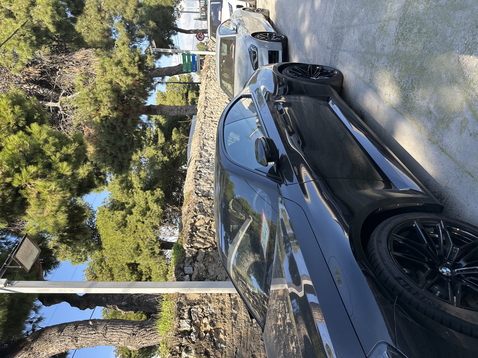 The image captures a sunny outdoor scene featuring two parked cars alongside a road, backed by a natural stone wall and dense green pine trees. The perspective is tilted, showing the scene from a slightly elevated, diagonal angle.
In the foreground, a glossy black BMW, possibly an M series sedan or coupe, is prominently parked with its side profile visible. It features large, multi-spoke black wheels, one of which clearly displays "Pirelli" brand text and tire specifications "295/35 R21". Reflections of the surrounding trees and sky are visible on the car's sleek surface. A small, partially visible sticker inside the car's window has "METAMORFOSI" printed on it, confirming the location context of Metamorfosi, Greece.
Behind the black car, a lighter grey or silver sedan, also appearing to be a BMW, is parked. Further back, on the opposite side of the road, more cars are partially visible.
The setting suggests a Mediterranean environment, characterized by the type of evergreen trees and the rustic stone wall lining the road. A clear blue sky is visible above the trees. A signpost with multiple green and blue directional panels stands in the mid-ground, though its text is illegible. A small red circular road sign with a white horizontal bar, indicating "No Entry," is also visible near the grey car.
The time of day is bright daylight, indicated by the strong illumination and visible shadows. No people are present in the image. The scene is static, focusing on the parked vehicles and their natural surroundings.
