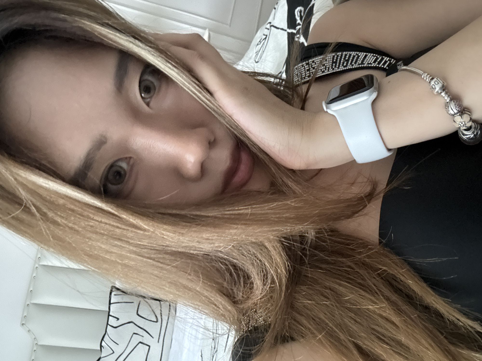 An individual is resting their head on their hand, looking towards the camera. They have long, light brown hair, brown eyes, and are wearing a black top with a sparkly strap. A white smartwatch and a charm bracelet are visible on their left wrist. In the background, there is a white upholstered headboard with decorative studs and pillows with a black and white graphic pattern on a bed. The lighting suggests an indoor setting, possibly a bedroom. The location is not determinable from the image alone.