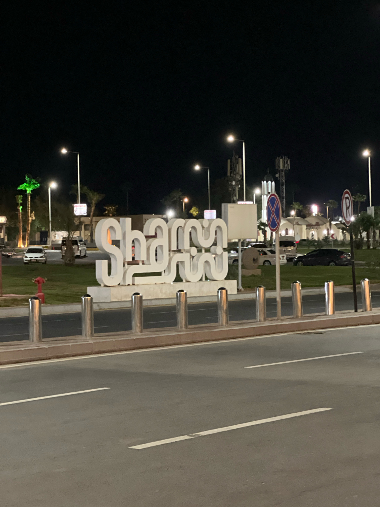The image shows a large, white, three-dimensional sign spelling out "Sharm" in Arabic script, illuminated at night. The sign is located in front of a grassy area with palm trees and streetlights in the background. Several cars are parked or driving in the vicinity, and some buildings, possibly in a Mediterranean architectural style, are visible. There are also traffic signs and a fire hydrant in the scene. The overall setting suggests an urban environment in Sharm El Sheikh, Egypt, likely a public space or roadside attraction.