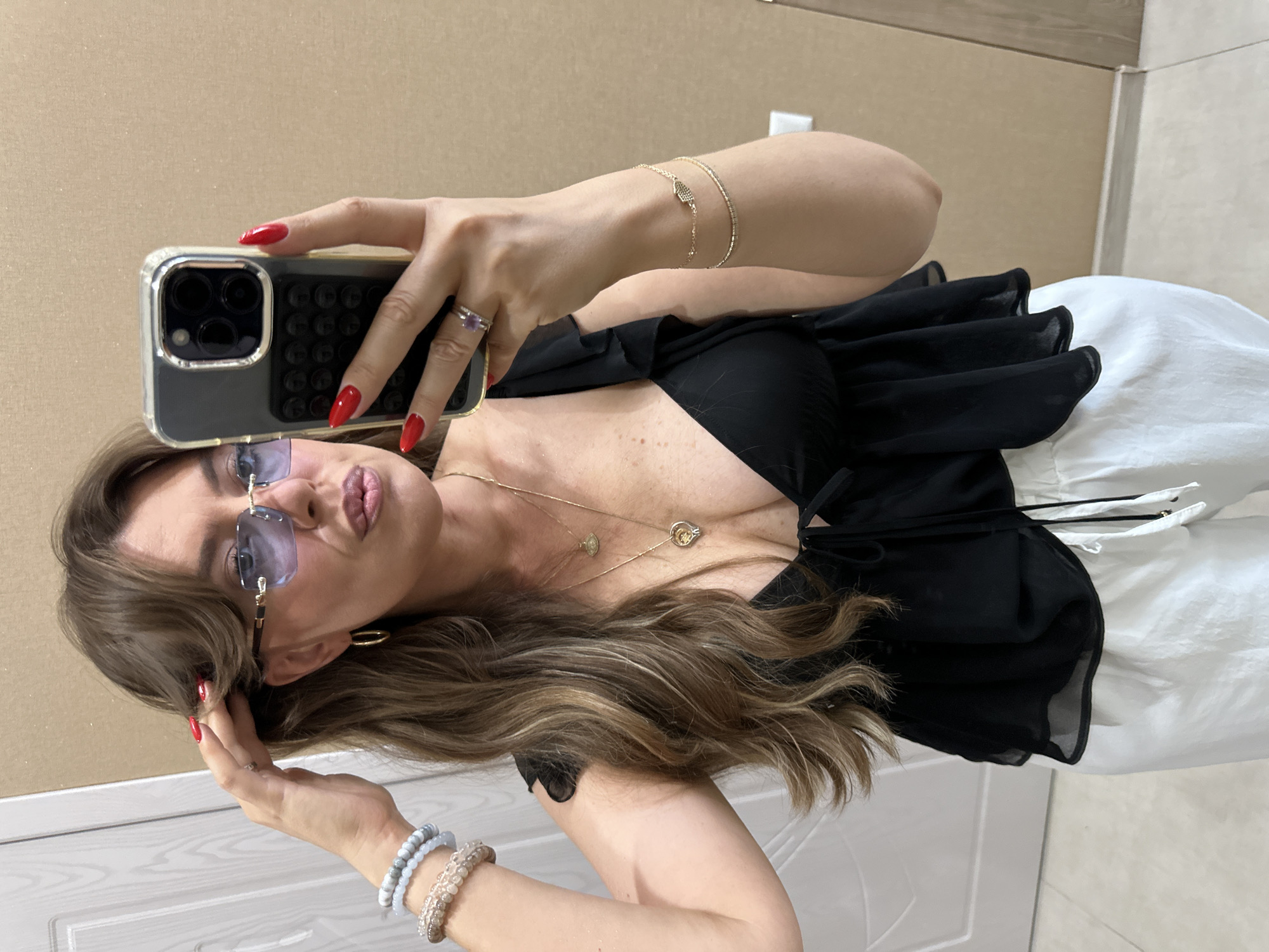 A woman is taking a mirror selfie in a room with beige walls. She has brown hair styled with bangs, is wearing light blue rimless glasses, and red nail polish. She is wearing a black top with ruffled sleeves and a V-neckline, and white pants. She is adorned with several bracelets on her left wrist, a necklace with a heart pendant, and a ring on her right hand. The background shows a white door on the left and a portion of the beige wall. The overall impression is a casual indoor setting.
