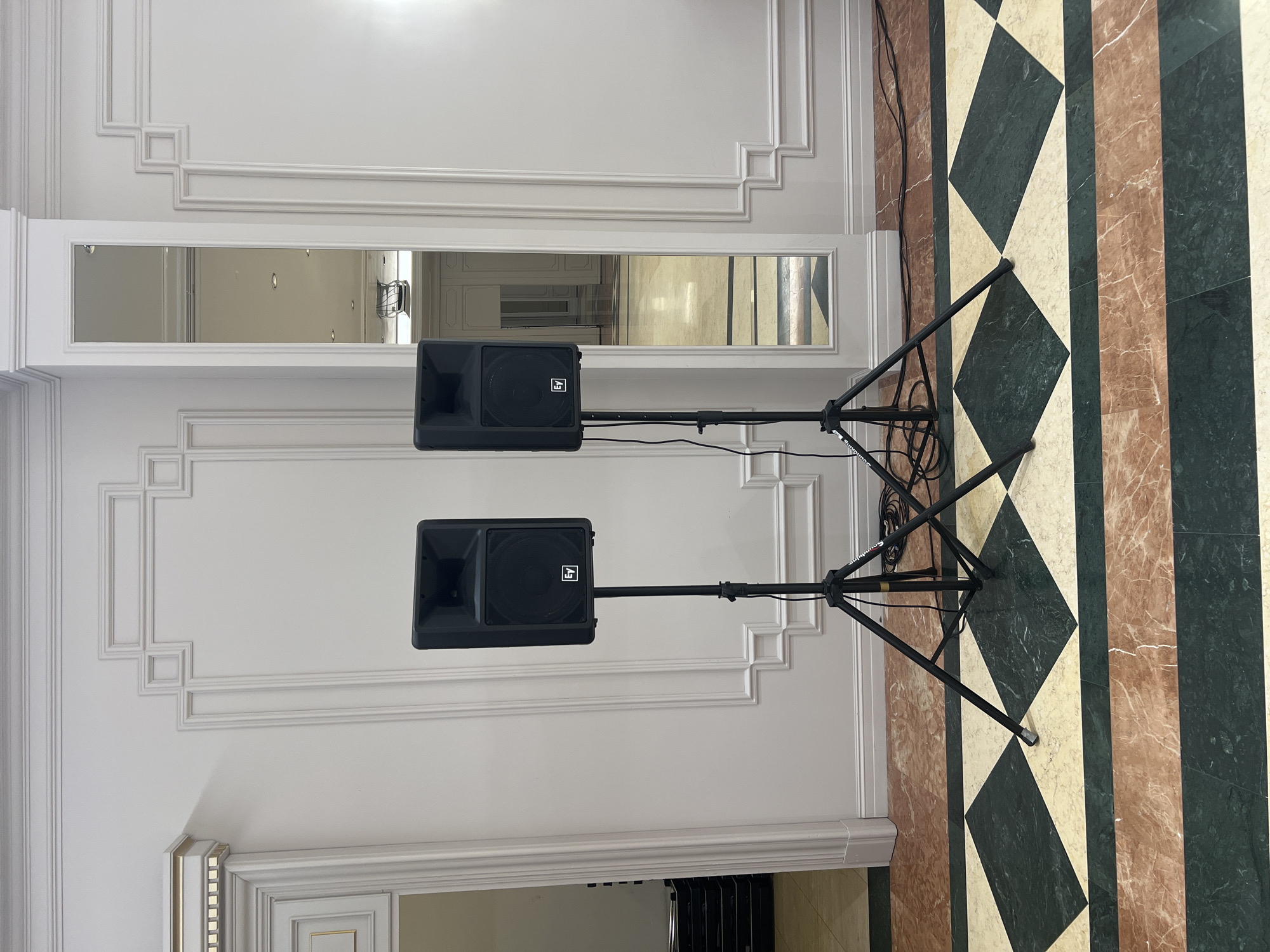 The image shows two black Electro-Voice brand speakers mounted on black tripod stands. The speakers are positioned centrally in a room with white walls featuring decorative molding. The floor is a marble mosaic with alternating dark green, beige, and pinkish-brown tiles. To the right, a tall mirror reflects the room, showing a hallway with more molding and a ceiling with recessed lighting. To the left, a doorway leads to another room with stacked black equipment. The lighting is bright, suggesting an indoor event space.