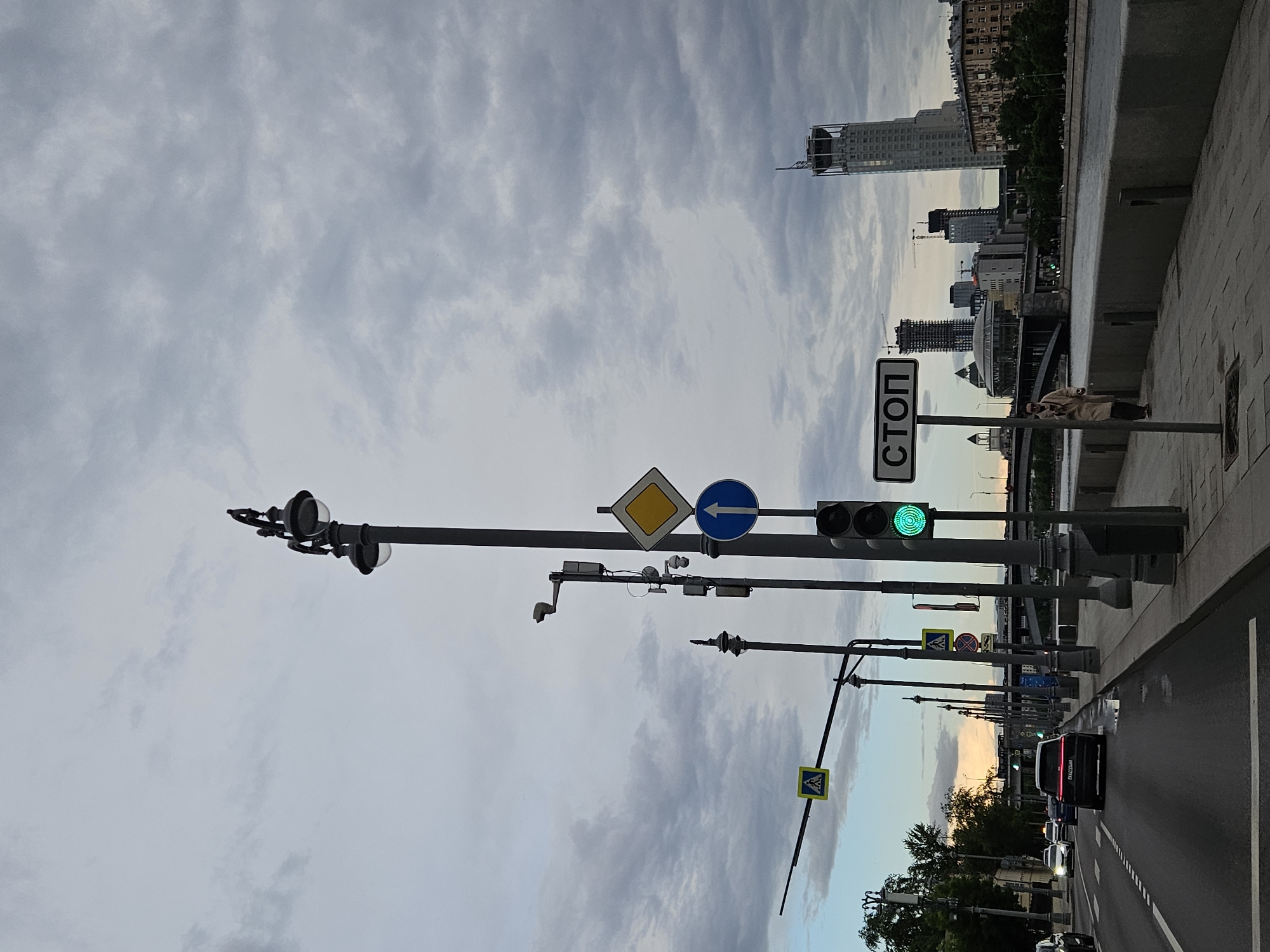 The image depicts a street scene in Moscow, Russia. The sky is overcast with gray clouds. A row of traffic signals is visible along the road. There are buildings in the distance, including a high-rise. A sidewalk runs alongside the road, and there is a person standing on it. Cars are driving on the road. Street signs are present. A stop sign is visible.