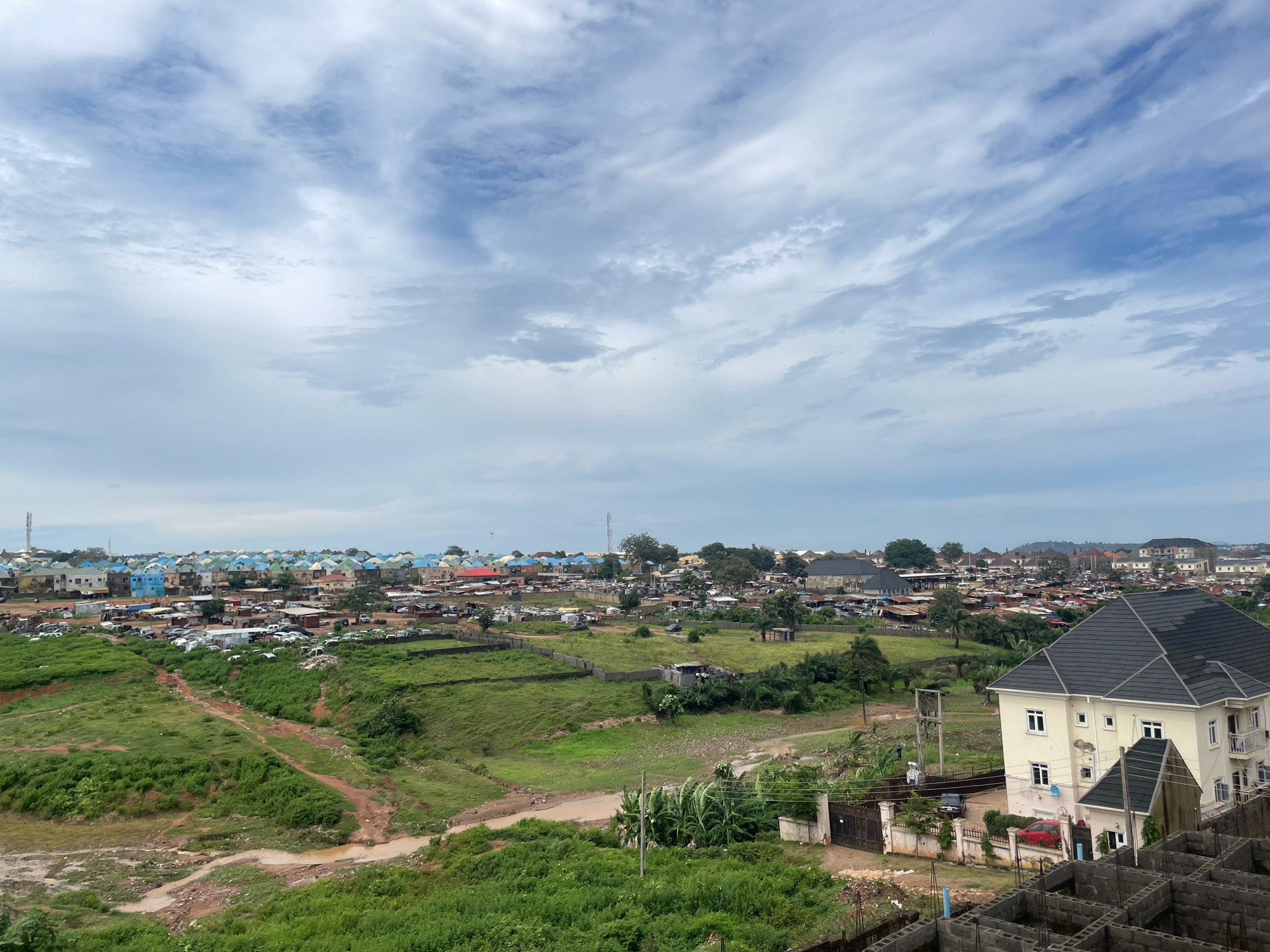 The image depicts an aerial view of a developing urban area in Abuja, Nigeria, characterized by a mix of residential buildings, open green spaces, and a cloudy sky. In the foreground, unfinished concrete structures suggest ongoing construction. To the right, a modern, multi-story building with a dark roof stands prominently, partially obscuring an older, multi-story structure behind it. A black gate and a red car are visible near the fence of the modern building.
The middle ground reveals a sprawling landscape of predominantly green terrain with scattered trees. Several dirt paths wind through the grassy areas, and a small, muddy stream is present. Beyond this, a densely populated residential area unfolds, featuring numerous houses with colorful roofs in shades of blue, green, red, and beige. A significant number of parked cars are visible throughout this section of the city, suggesting a built-up community.
In the background, more buildings and structures extend towards the horizon, blending into the hazy, cloudy sky. The overall scene conveys a sense of expansion and development within an urban environment under a partially overcast sky. There are no distinct individuals or animals clearly visible in the image.
