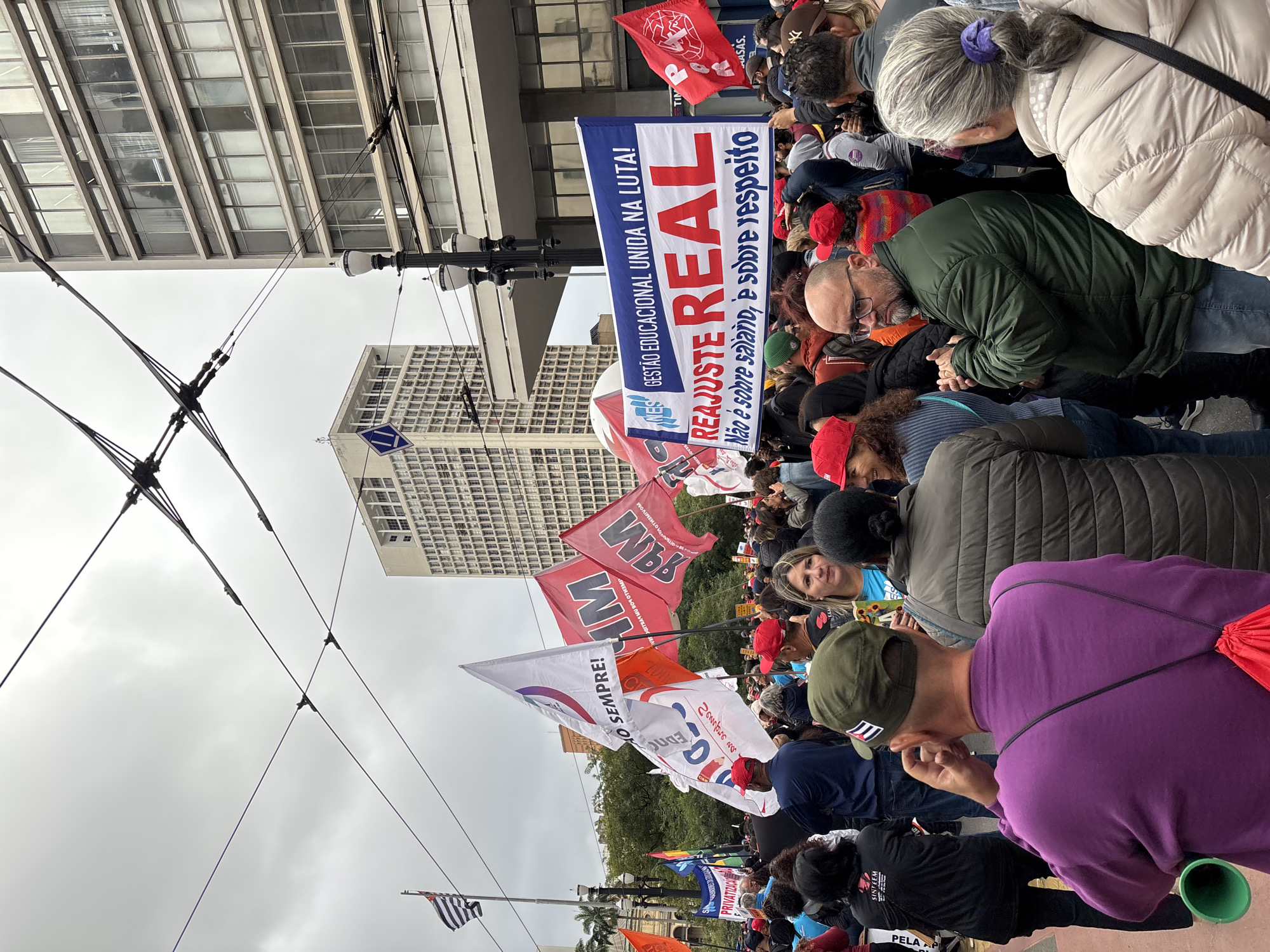 A large group of people is demonstrating outdoors in São Paulo, Brazil. The crowd is dense, and many individuals are holding signs and flags, some of which are in Portuguese. One banner reads "REAJUSTE REAL," meaning "REAL ADJUSTMENT" with the statement, "Não é sobre salário, é sobre respeito," "It's not about salary, it's about respect." Some flags are political. Overhead electrical lines are visible against a cloudy sky, with tall buildings in the background. One building bears a blue and white logo. The overall scene suggests a political rally or protest.