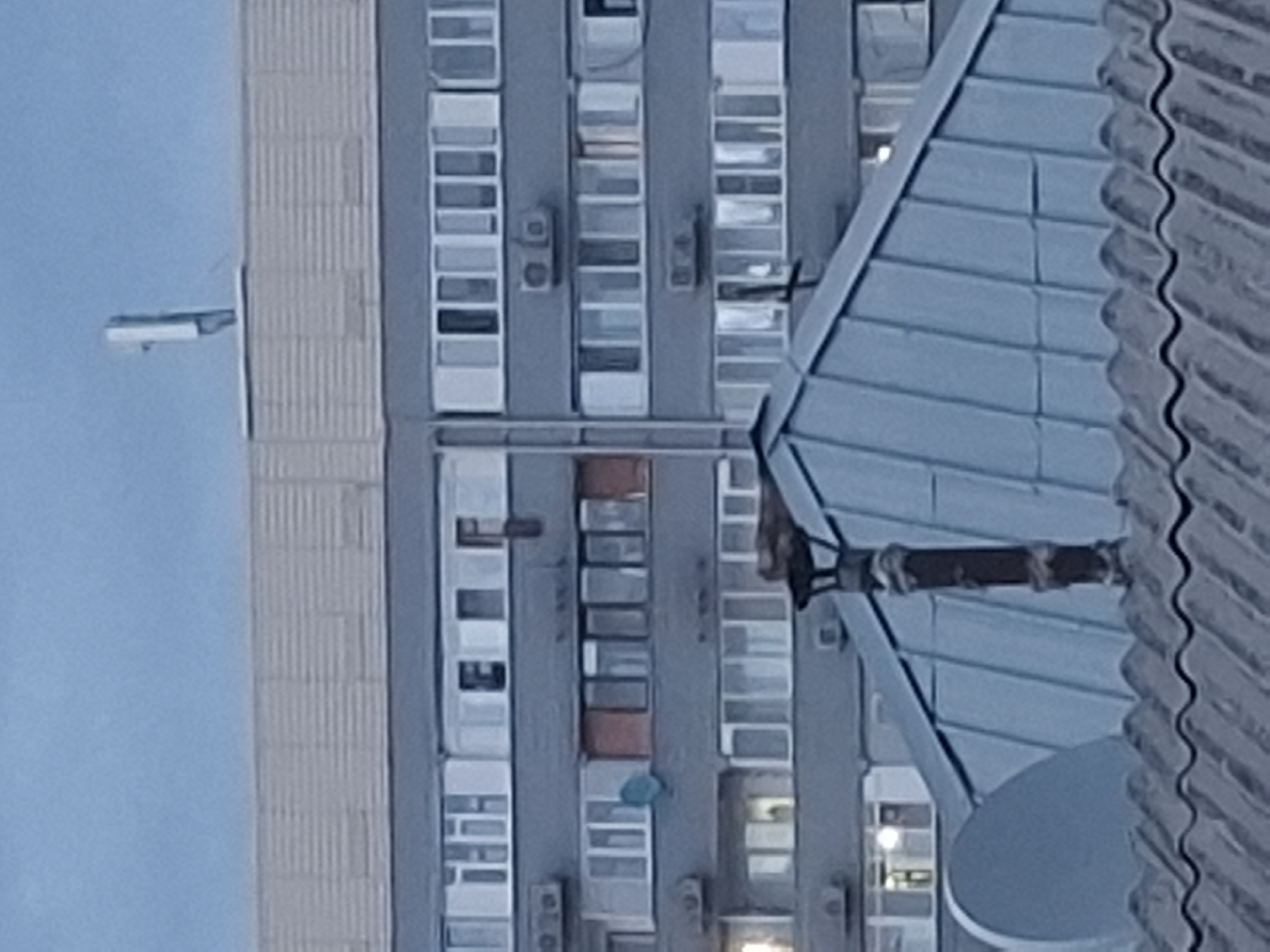 The image shows a close-up view of apartment buildings in Almaty, Kazakhstan. The buildings are multi-story and appear to be constructed from concrete and brick. The foreground features a corrugated metal roof with a satellite dish and a chimney. The middle ground shows a gray apartment building with numerous windows and air conditioning units. In the background, a taller, lighter-colored building is visible, topped with what appears to be an antenna. The sky is overcast and a pale blue, suggesting either early morning or late evening. No people are visible in the image.