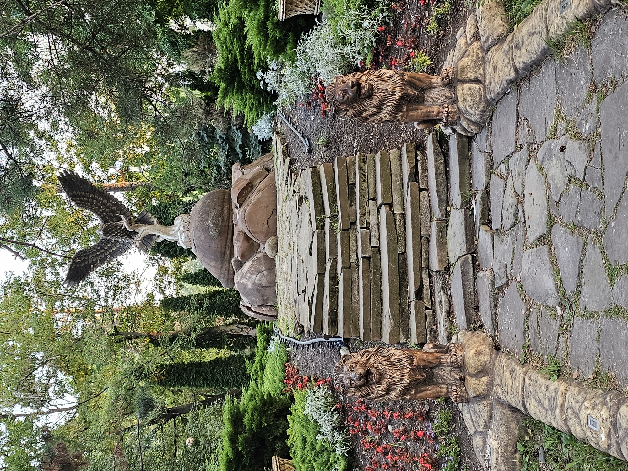 The image shows a stone pathway leading up a flight of uneven stone steps. Flanking the beginning of the path are two lion statues. At the top of the steps, on a large rock formation, is a statue of an eagle with its wings spread. The pathway and steps are surrounded by lush greenery, including bushes, trees, and small red flowers. The overall scene is that of an outdoor monument or garden feature. This monument is located in the city of Cherkessk, Russia.