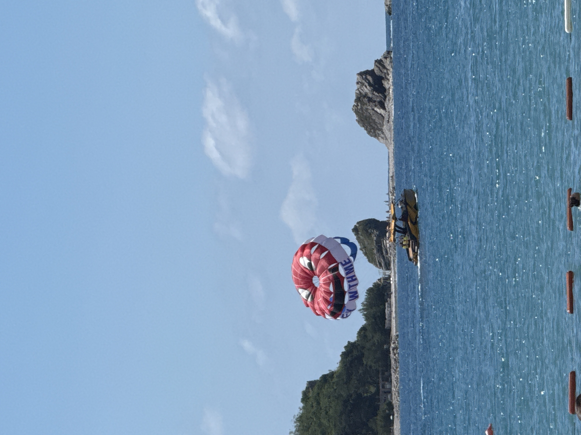 The image captures a vibrant daytime scene of parasailing over the clear blue waters off the coast of Budva, Montenegro. In the mid-ground, a large, colorful parasail, predominantly red and white with blue accents, is fully inflated and suspended in the bright blue sky. On its blue segment, the partial text "WTHME" is visible.
Below the parasail, on the sparkling sea, a boat, likely the one towing the parasail, is partially visible towards the right side of the frame. The water is a brilliant turquoise-blue, reflecting sunlight with numerous bright glints. The background features a rugged coastline with dark, rocky cliffs and hills densely covered in green trees, indicating a lush, natural environment. A few soft, white clouds are scattered across the otherwise clear blue sky. The overall impression is one of a serene yet active recreational setting on a beautiful, sunny day. No people are distinctly visible, but their presence is implied by the parasailing activity.