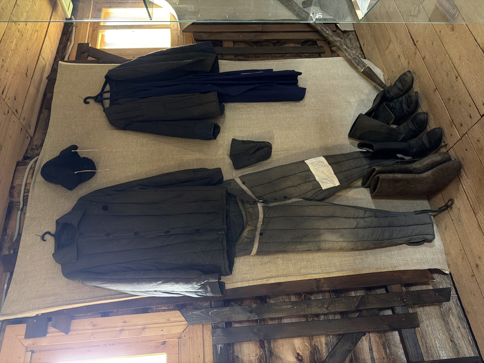 The image depicts a display of historical workwear, likely in a museum setting, located in Ust'-Pyzas, Russia. The clothing items, consisting of a quilted jacket, trousers with white banding, a dark hat, and several pairs of boots, are arranged on a draped fabric backdrop against a wooden wall. The garments are displayed on hangers or laid out on the floor. The environment suggests an indoor space with wooden walls and flooring, illuminated by natural light from windows. The display appears to be part of a cultural exhibition documenting the region's history or occupational traditions.
