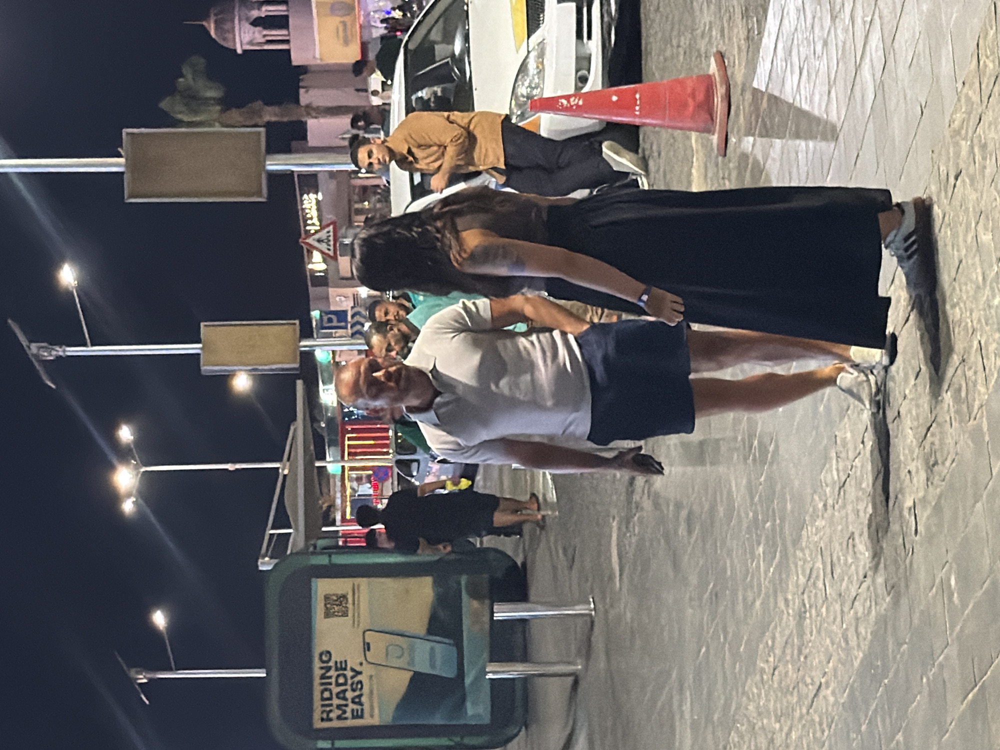 The image shows a night scene on a paved walkway in Sharm El Sheikh, Egypt. Two people are the primary subjects: a man in a light-colored polo shirt and dark shorts, and a woman wearing a dark, long skirt and a dark top. They appear to be standing and talking. In the background, there are streetlights, a white car with a yellow stripe, a red traffic cone, and various buildings and signs suggesting a commercial or entertainment area. A large green advertisement sign is visible on the left, displaying the text "RIDING MADE EASY." Other individuals are present in the background, moving around. The lighting indicates it is after dark, with artificial lights illuminating the scene.